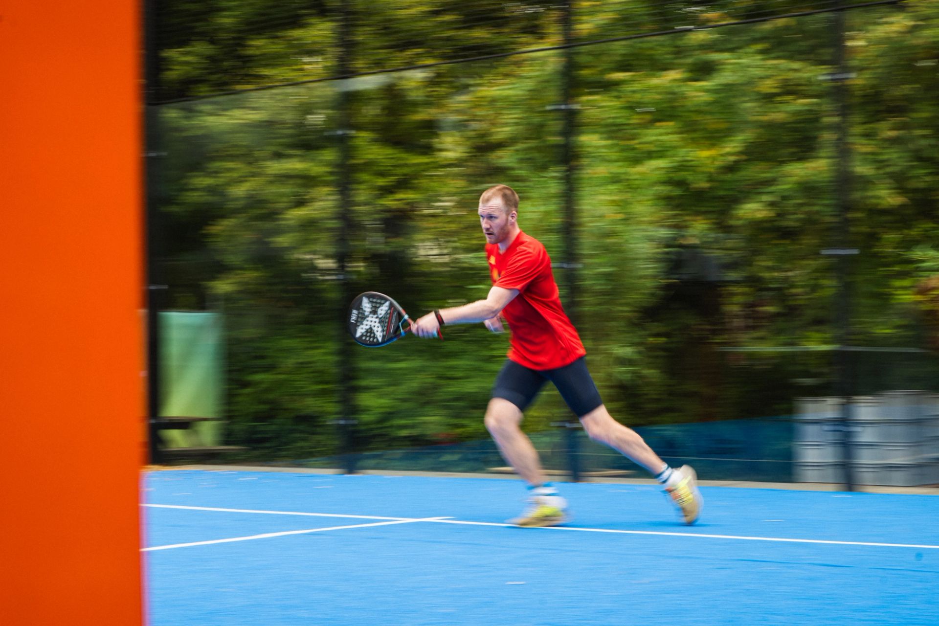 A padel player in a red shirt lunges across a blue outdoor court to return the ball.