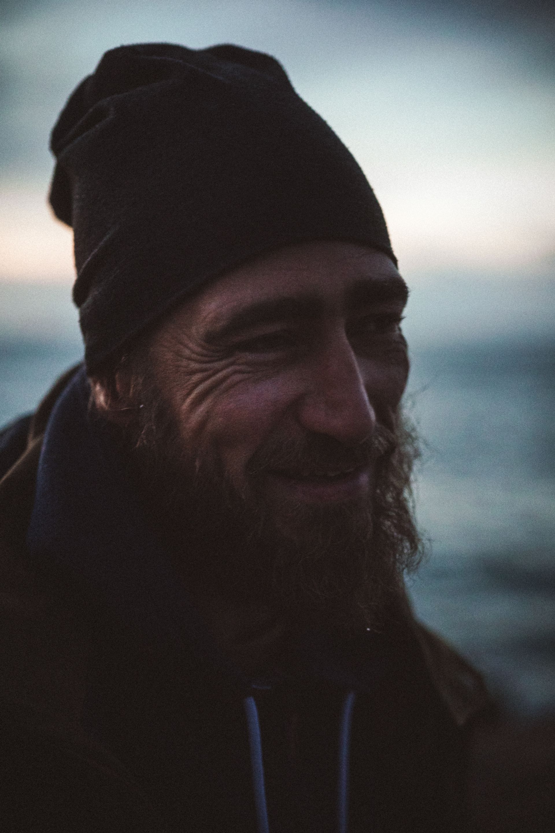 A man with a beard and beanie smiles softly, with a blurred ocean background and dim lighting.