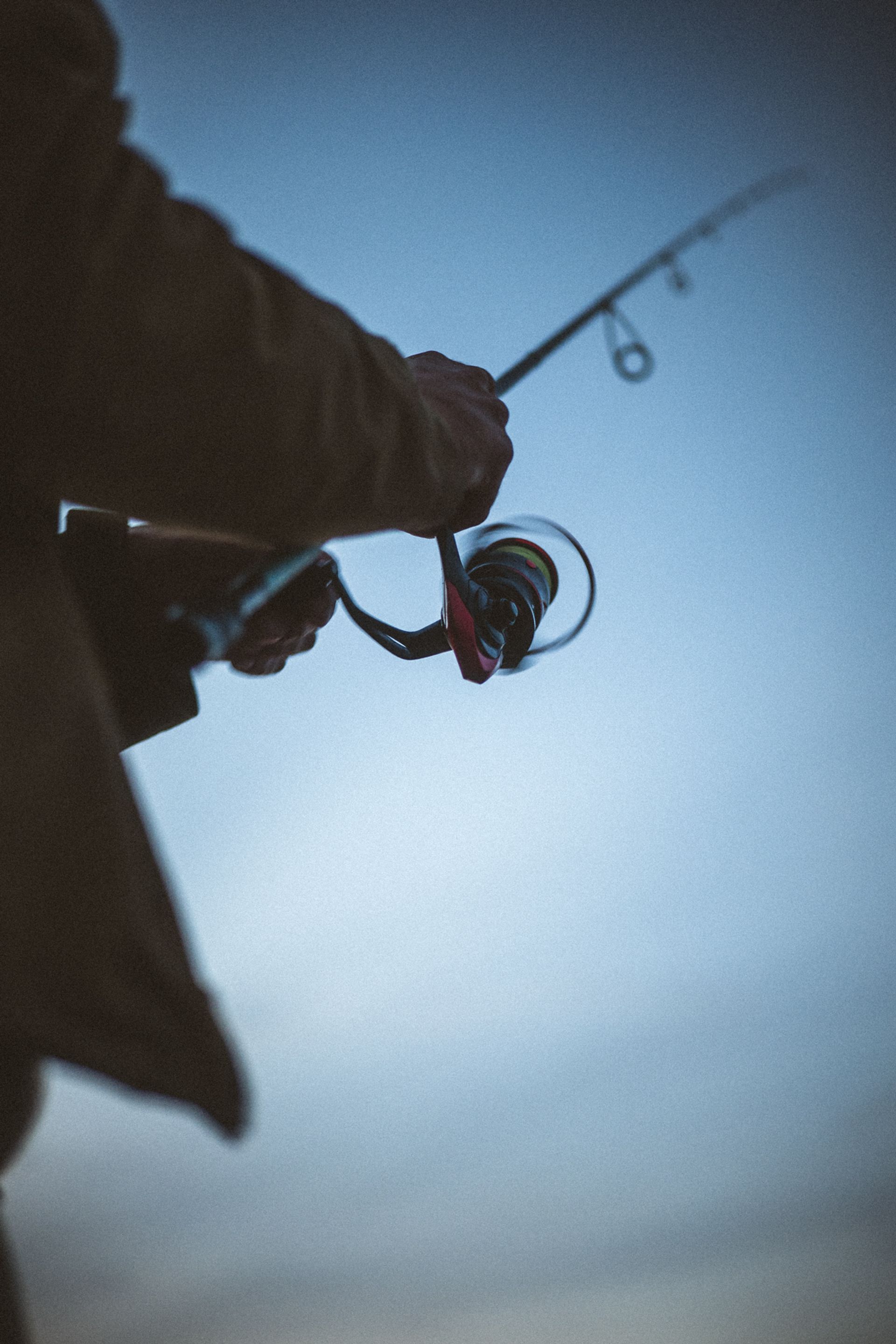 A close-up of a person's hand gripping a fishing rod, with a cloudy blue sky in the background.