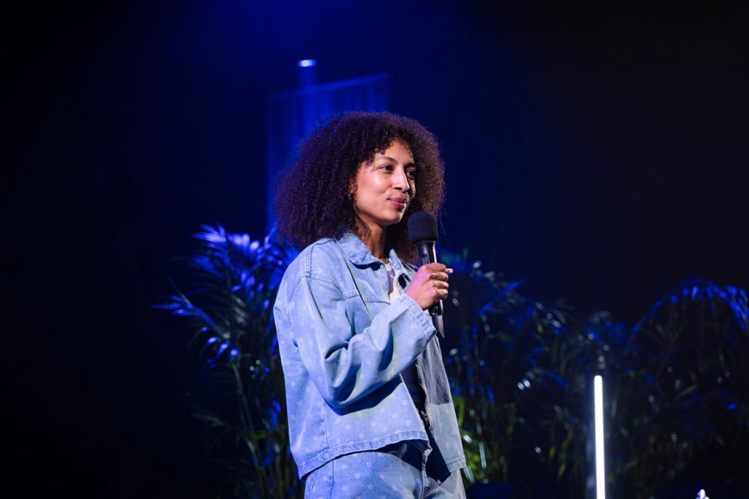 Une femme aux cheveux boucles en veste en jean clair parle au micro sur scene, eclairee en bleu avec des palmiers derriere.