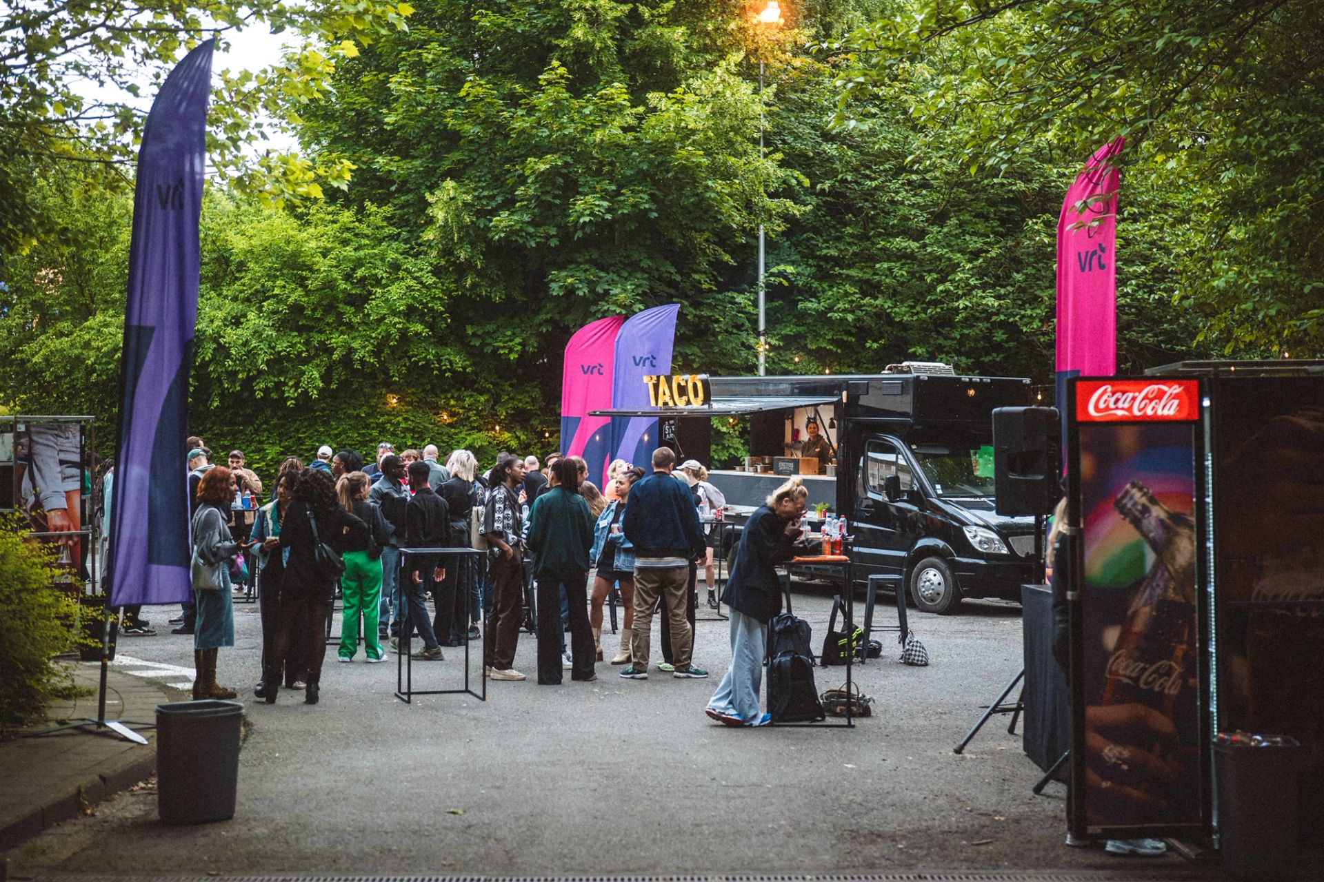 People gather around a food truck and VRT flags at an outdoor evening event among green trees.