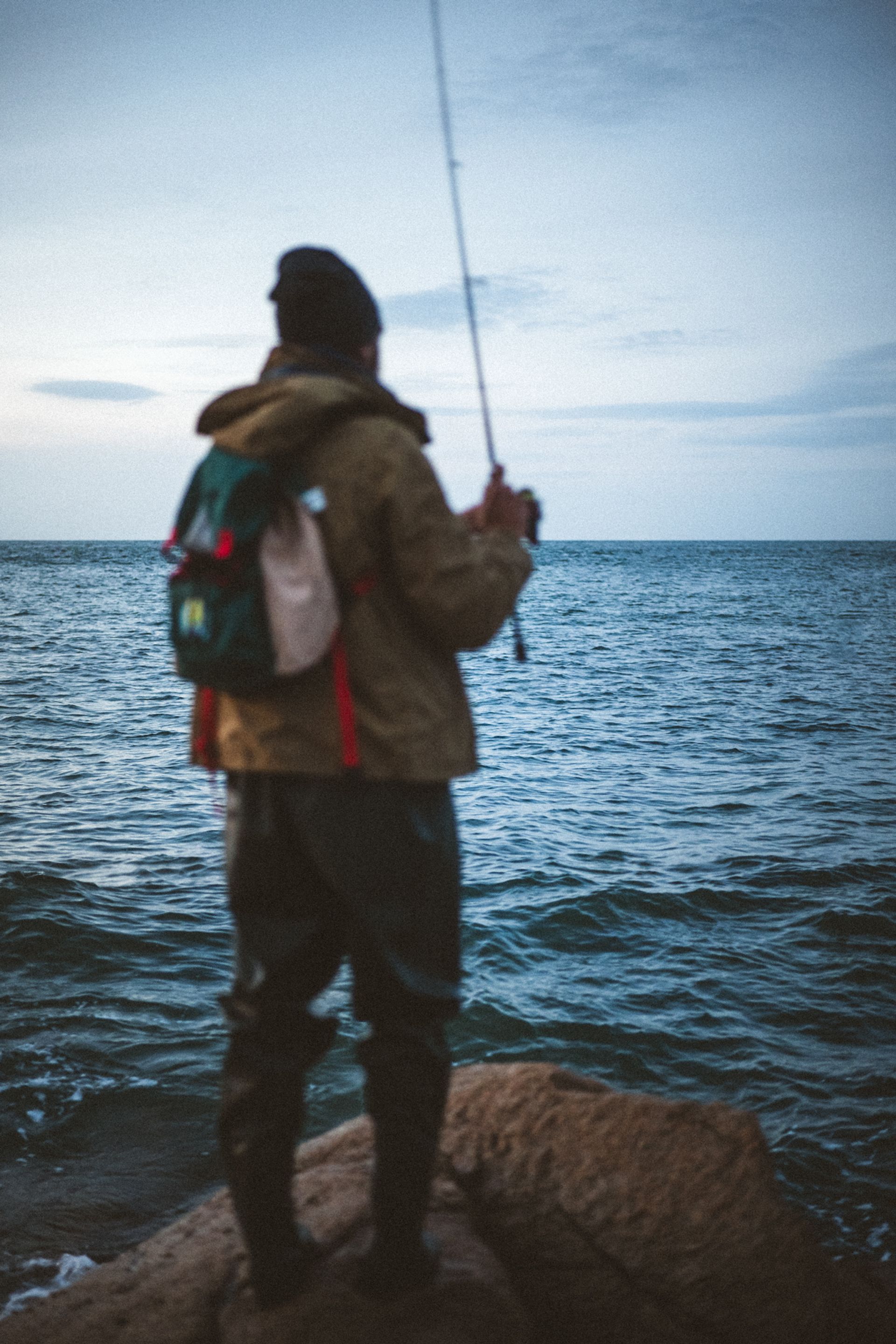 A person stands on a rock by the sea, holding a fishing rod, wearing a backpack and fishing attire.