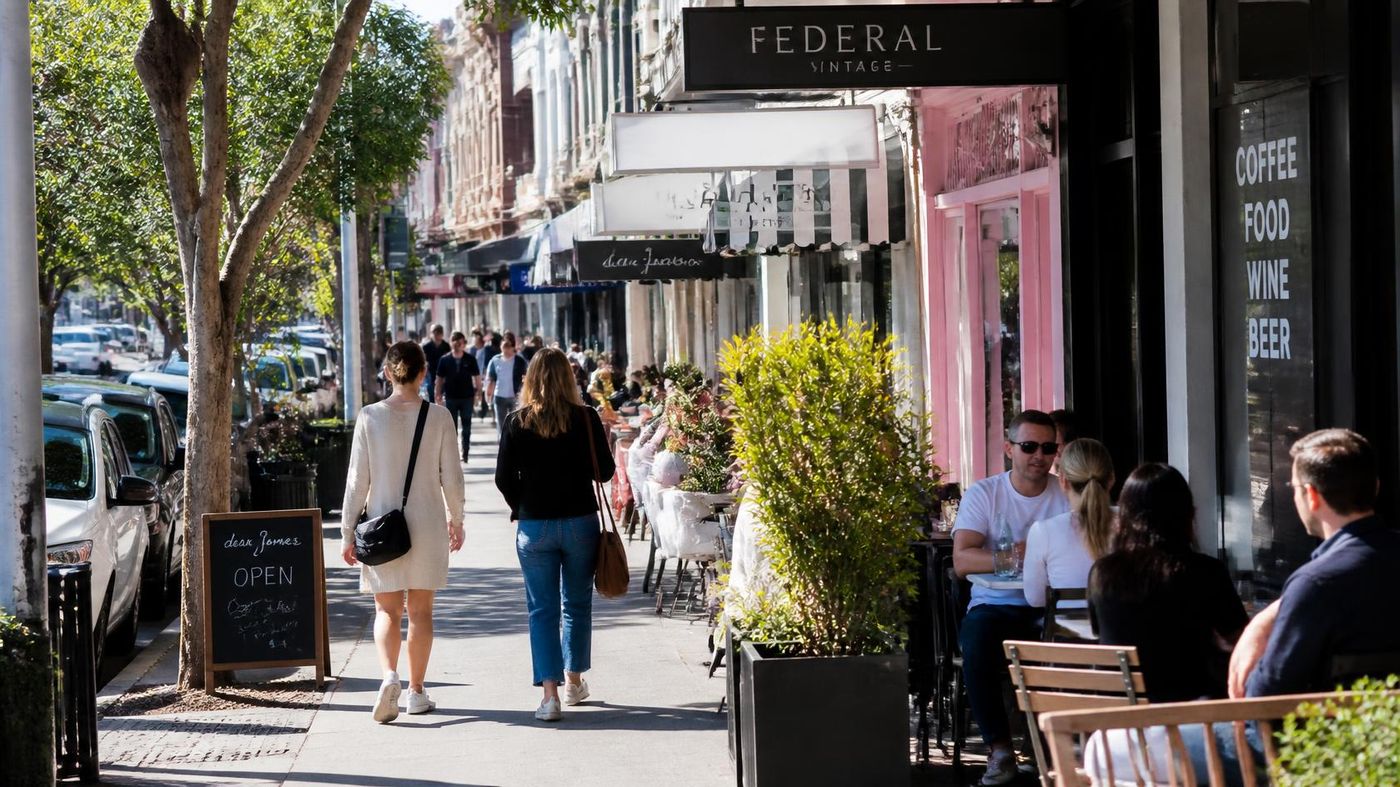 Pedestrians walking past cafes and boutique shopfronts on a tree-lined city street, with outdoor dining tables and a 'Federal Vintage' shop sign.