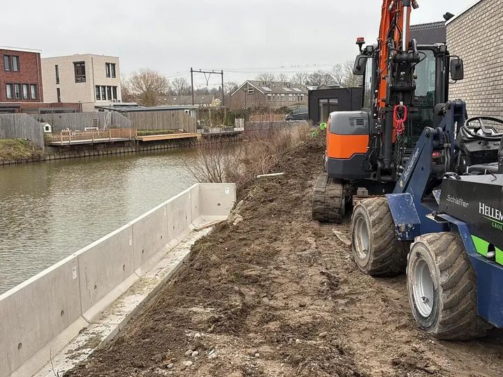 Een minigraver en de eigen shovel met de belettering Hellemons Groen naast een nieuwe betonnen keerwand langs het water