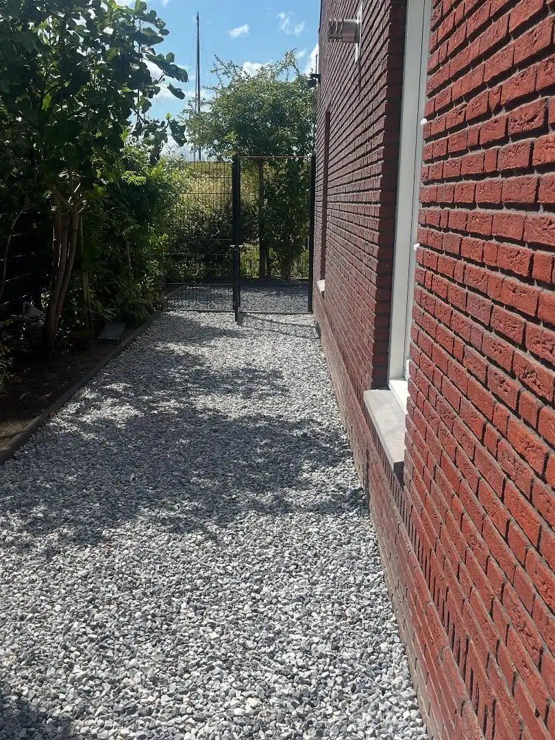 Zijpad van lichtgrijs grind langs een rode bakstenen gevel, met struiken aan de andere kant