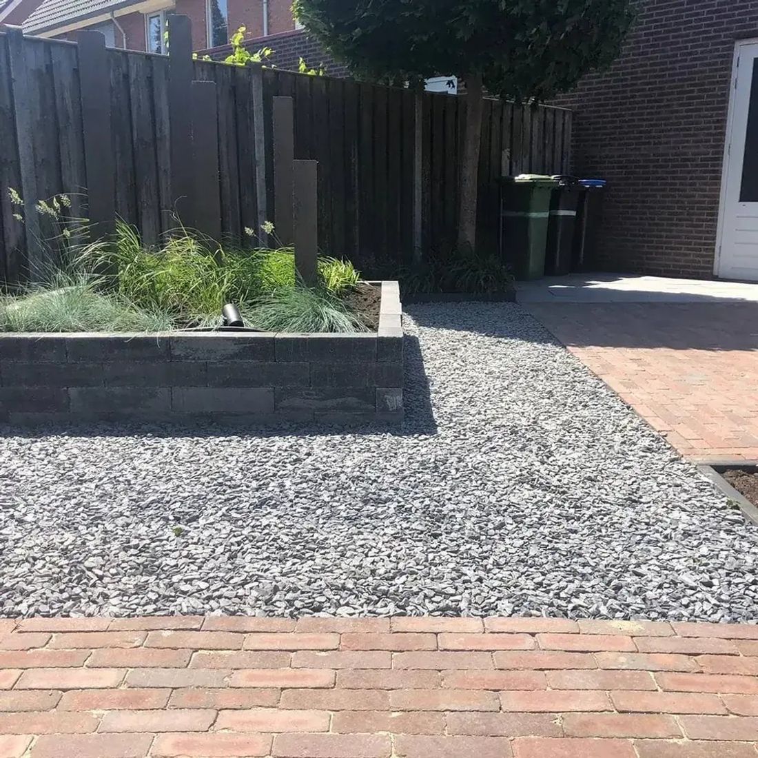 Verhoogde plantenbak van donkergrijze stapelblokken met siergrassen, grijs split en een pad van rode klinkers