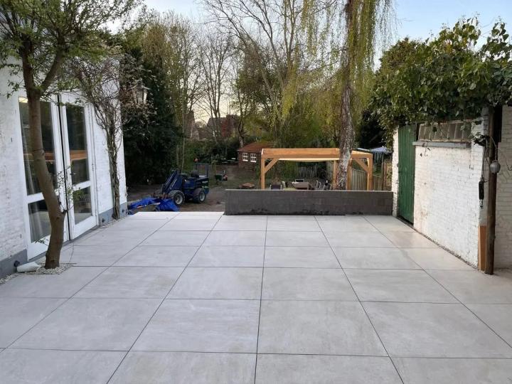 Groot terras van lichtgrijze keramische tegels achter een wit huis, met achterin een lage muur en een houten pergola