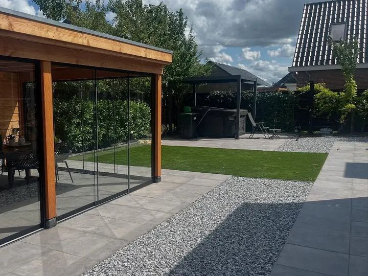 Achtertuin met een houten overkapping met glazen schuifwanden, grote grijze tegels, een strook wit grind en kunstgras