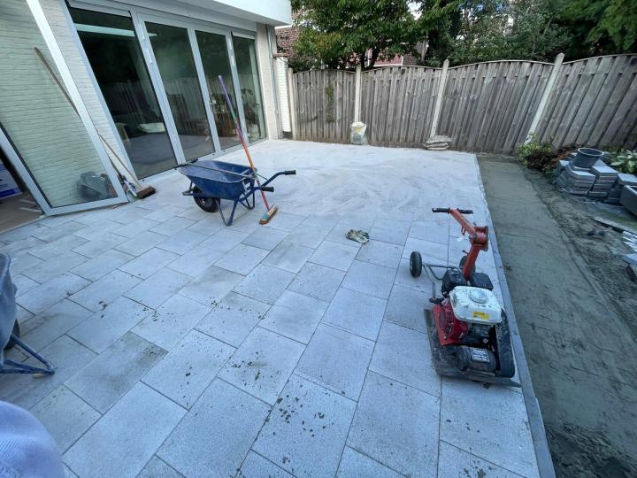 Lichtgrijs tegelterras in aanleg achter een woning, met kruiwagen en trilplaat