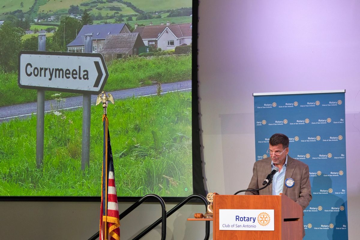 Mark Figart speaks at the podium beside a projected slide showing a roadside sign for Corrymeela in the green Northern Ireland countryside.