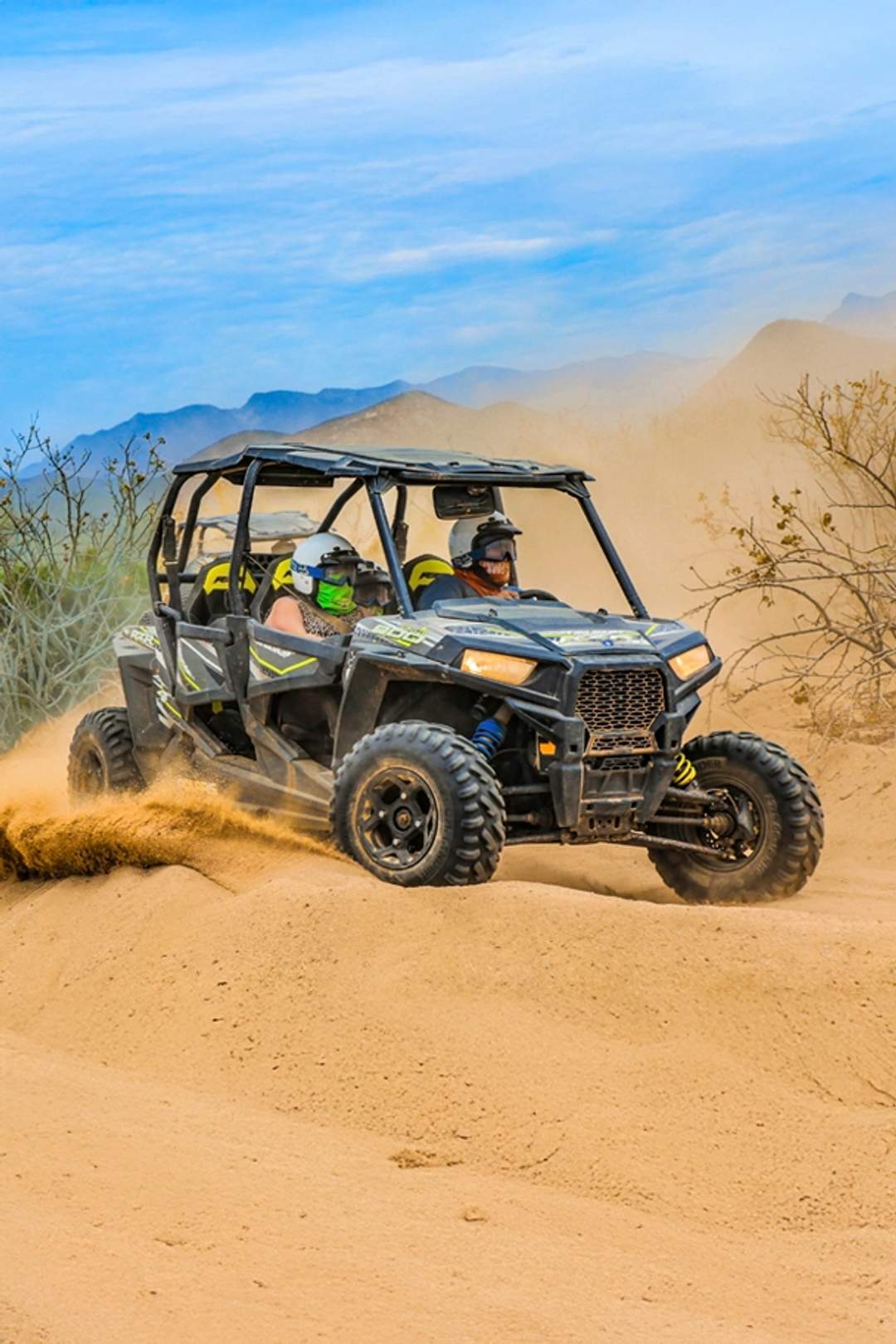 Two people drive a UTV on a sandy desert trail, kicking up dust as they navigate terrain with mountains in the background.