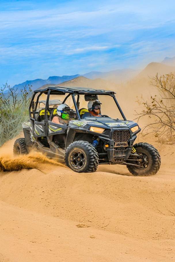 Dos personas en un UTV por un sendero en el desierto, levantando polvo mientras recorren un terreno con montañas al fondo.