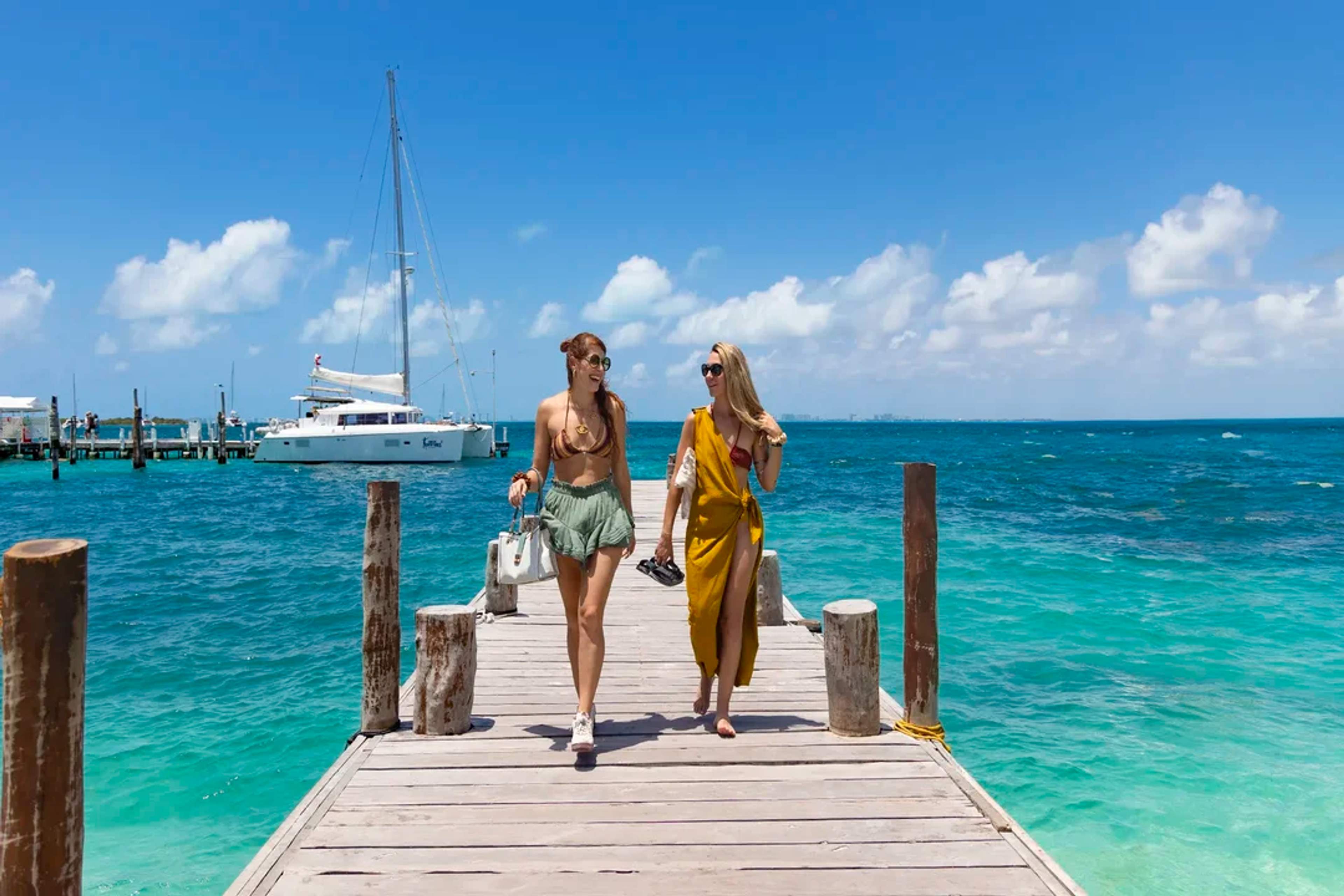 Two women walk on a sunny dock by turquoise waters with a catamaran in the background.