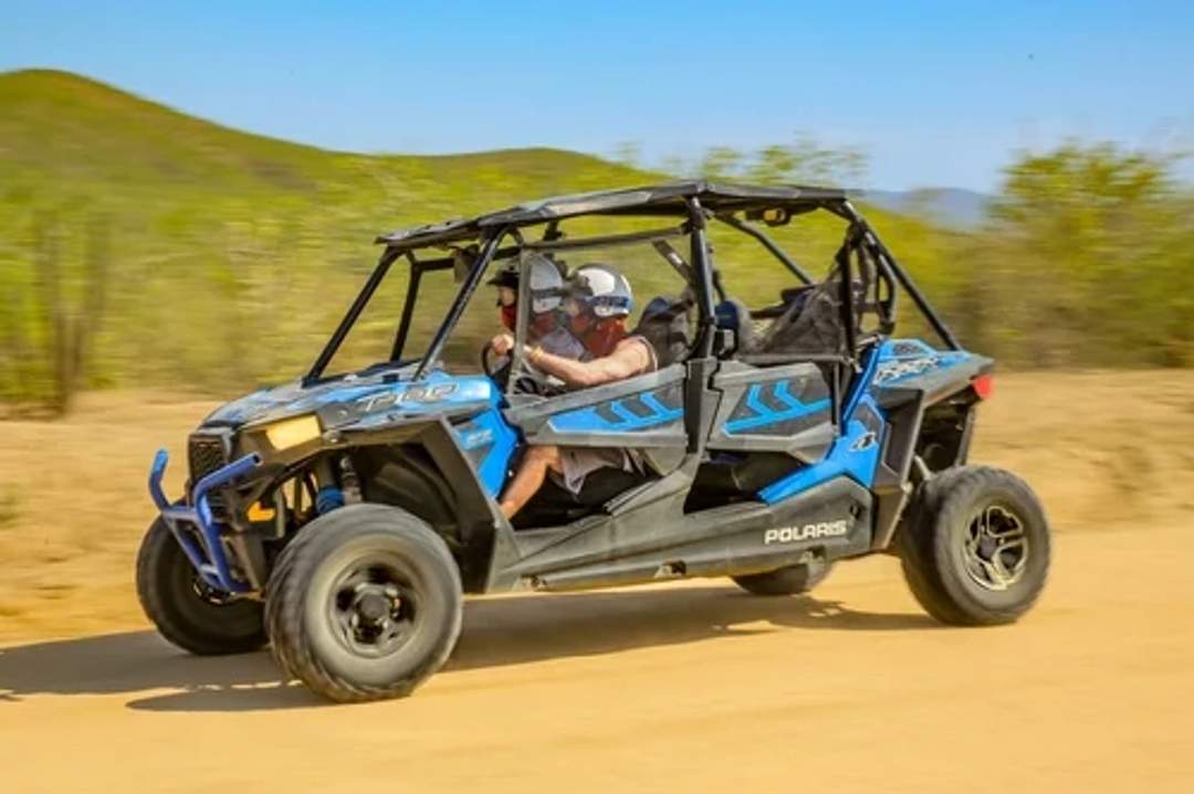Dos personas se divierten conduciendo un Polaris RZR a través del paisaje desértico de Cabo, con exuberante vegetación y montañas al fondo.