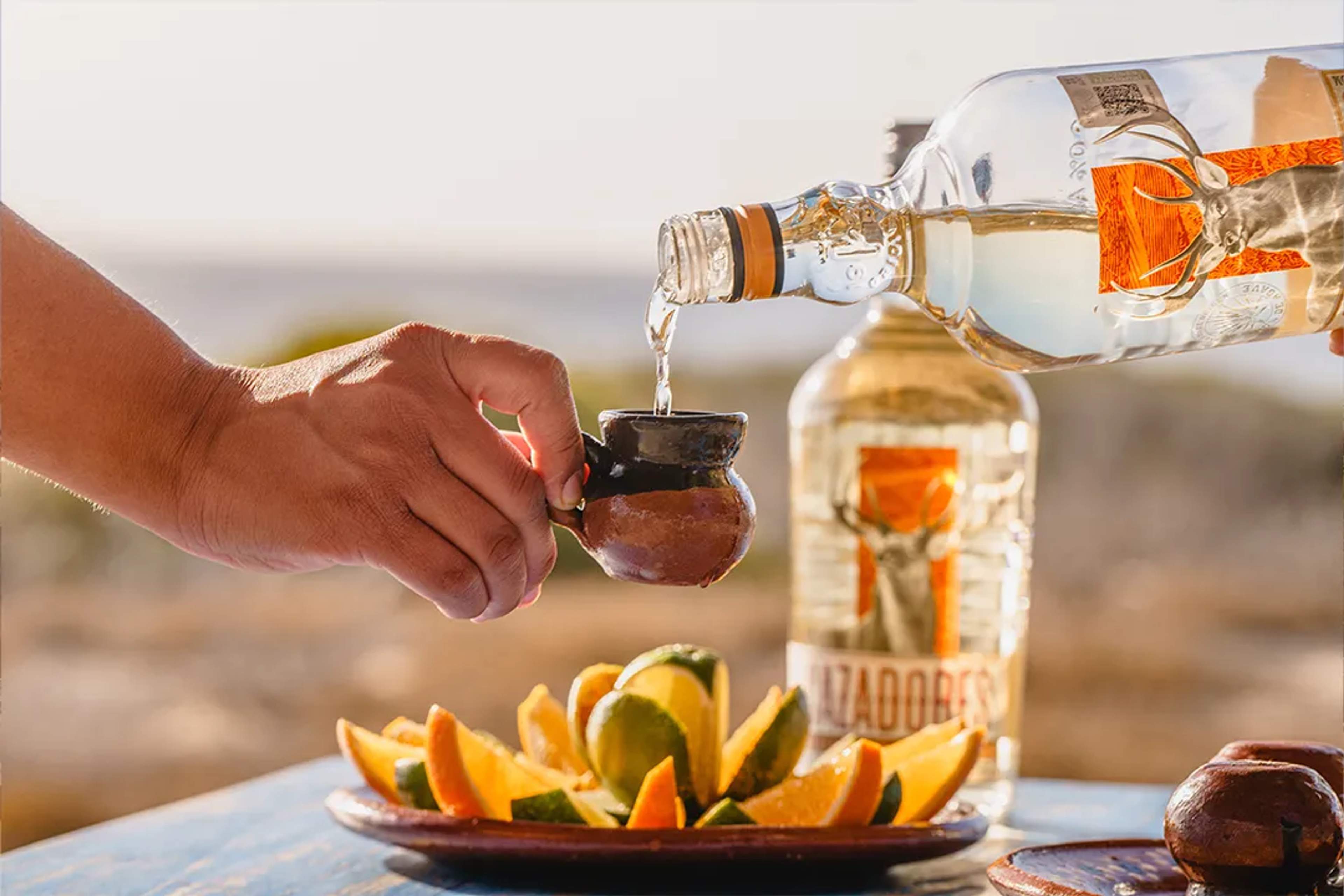 Tequila is poured into a clay cup beside a plate of lime and orange slices.