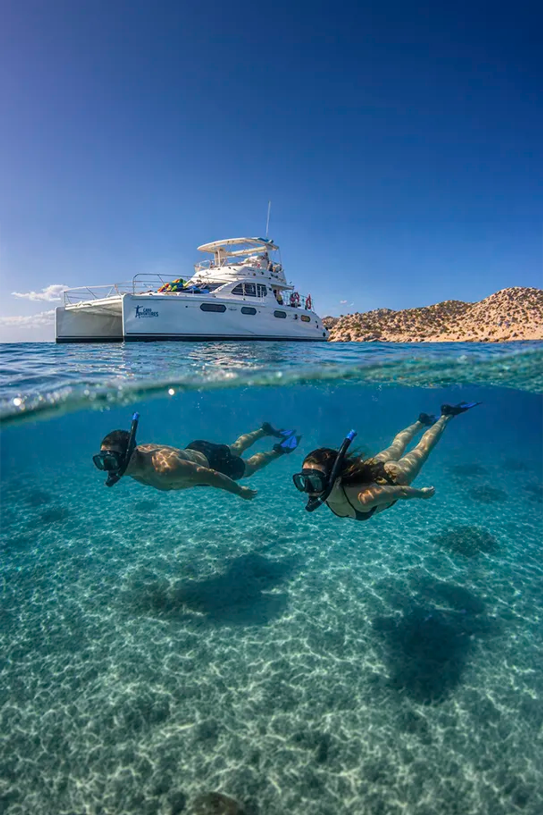 Couple snorkeling in clear turquoise waters below luxury catamaran near Cabo San Lucas desert coastline, Los Cabos Mexico