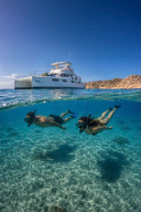 Pareja haciendo snorkel en aguas turquesa bajo catamarán de lujo frente a la costa desértica de Cabo San Lucas, Los Cabos