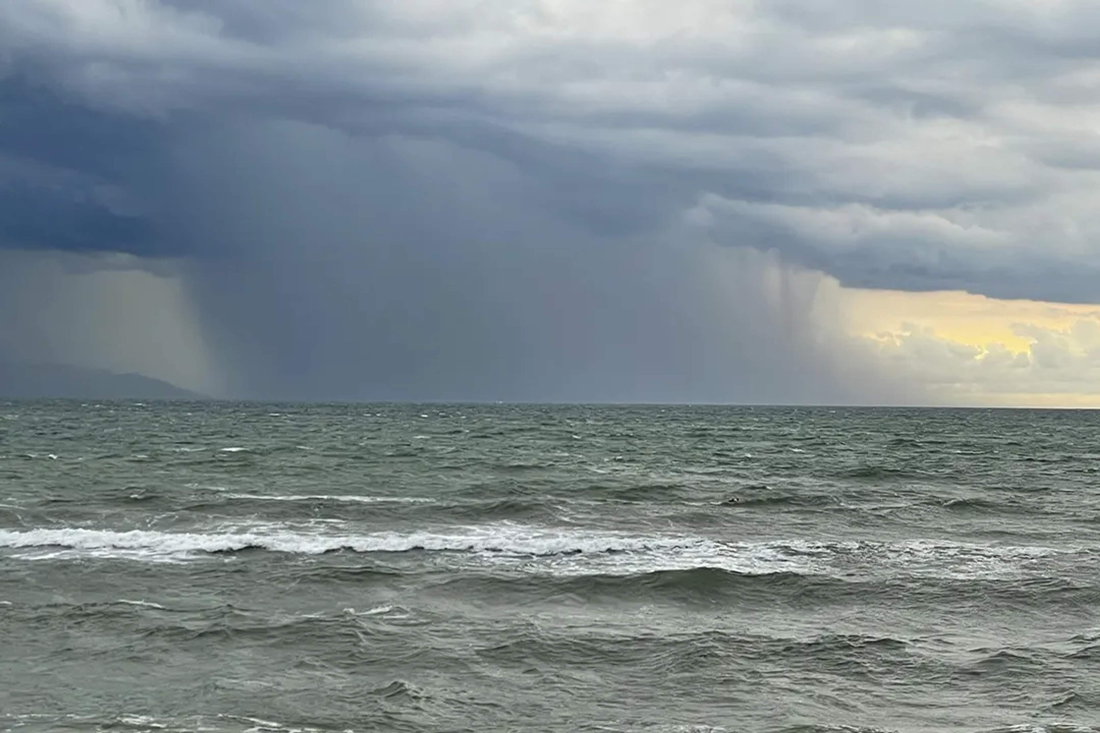 Dark storm clouds release rain over the ocean, while sunlight breaks through on the horizon.