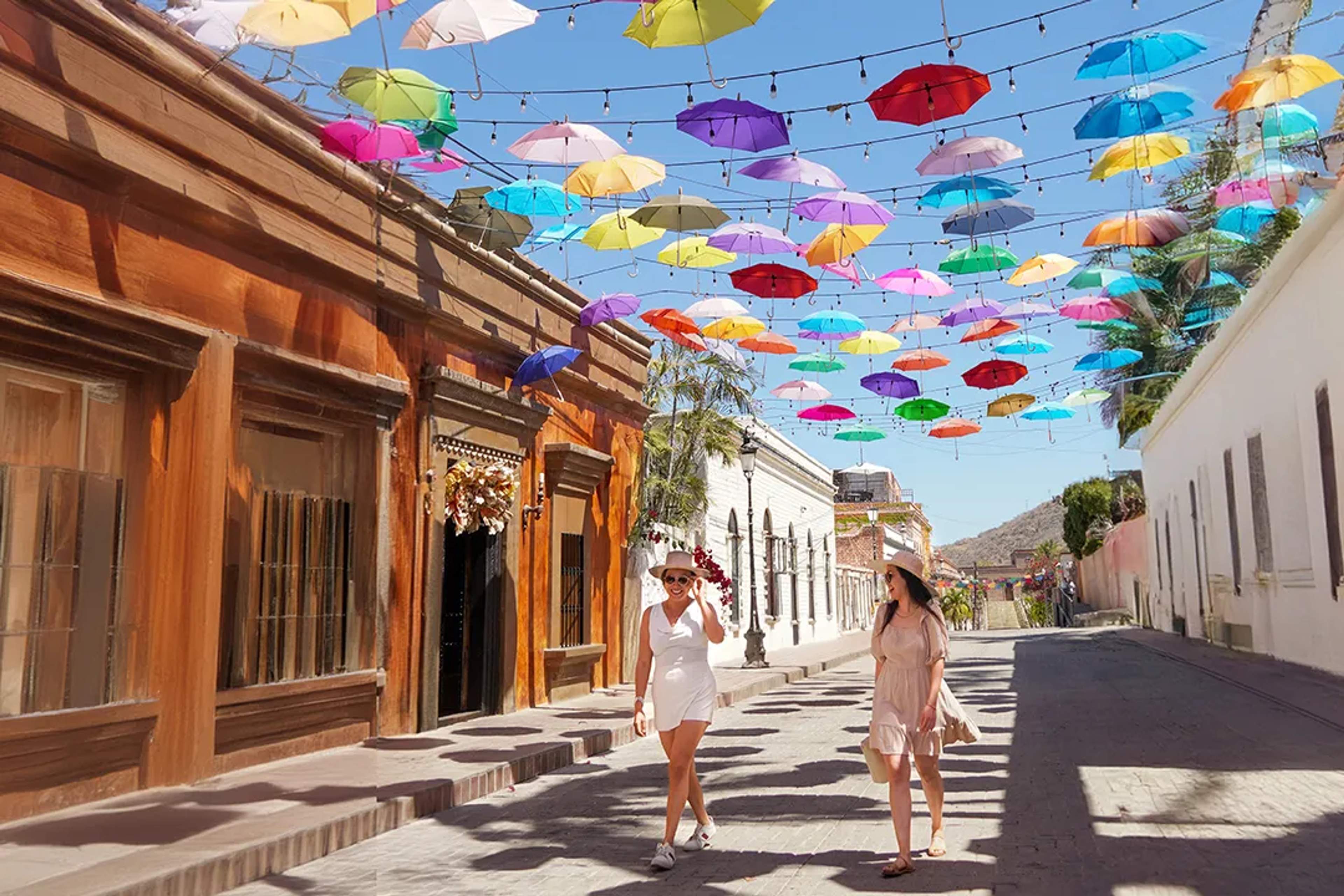 Dos mujeres caminan bajo coloridos paraguas colgantes en una calle empedrada de Todos Santos.