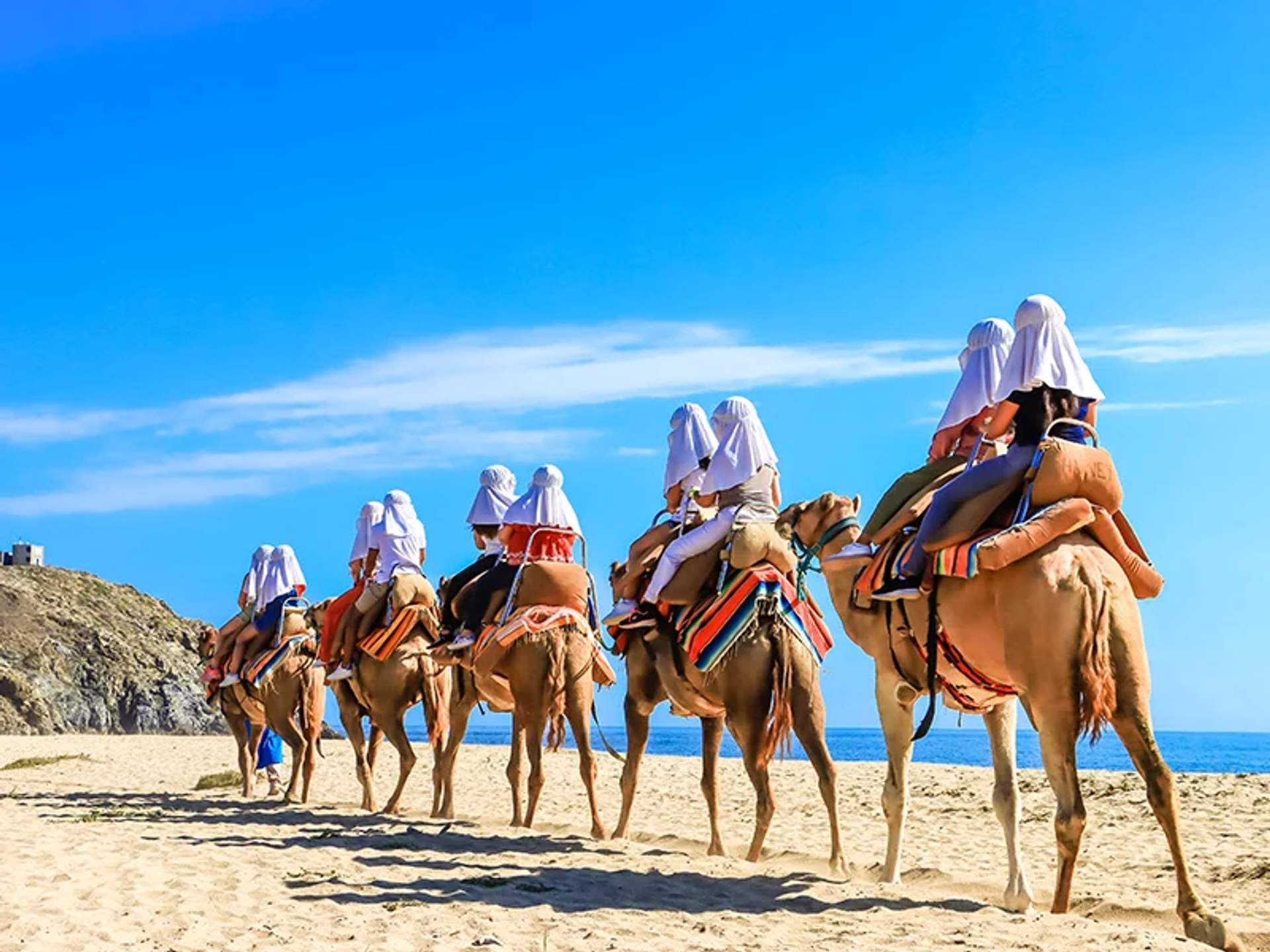 A group of people riding camels along a sandy beach under a clear blue sky.