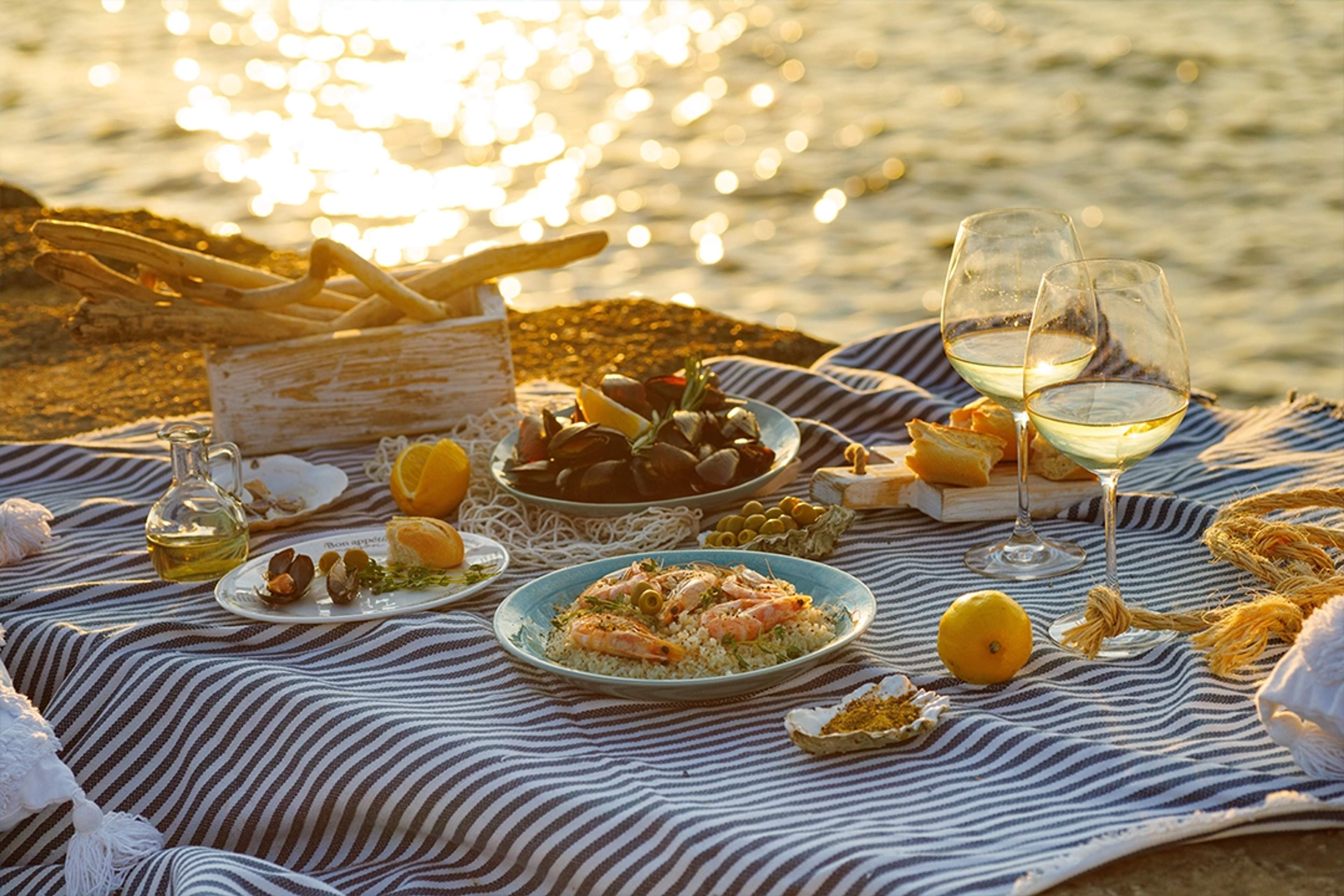 Un picnic junto al mar al atardecer con mariscos, pan, copas de vino y frutas cítricas sobre una manta a rayas.