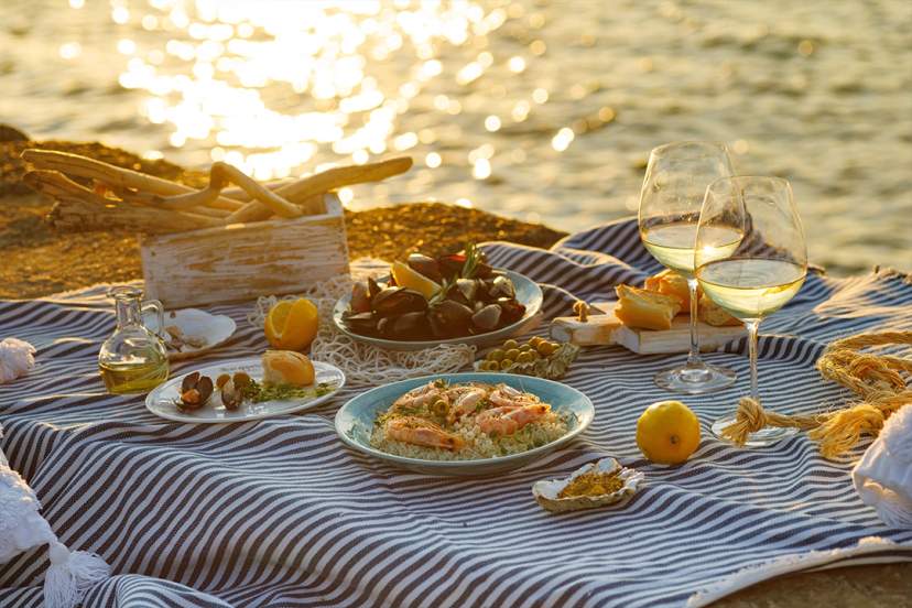 A seaside picnic at sunset featuring seafood, bread, wine glasses, and citrus fruits on a striped blanket.