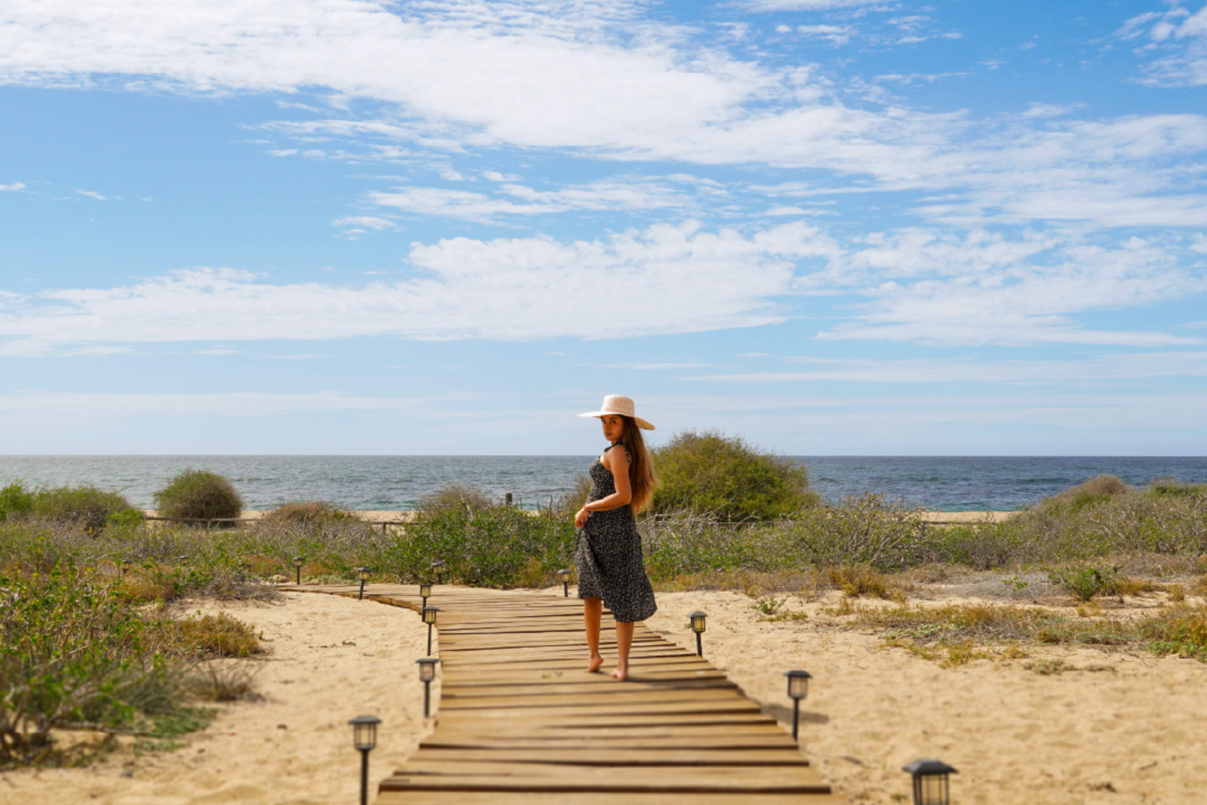 Woman in hat walking on wooden path to the ocean at Tierra Sagrada, Instagram spot in Cabo