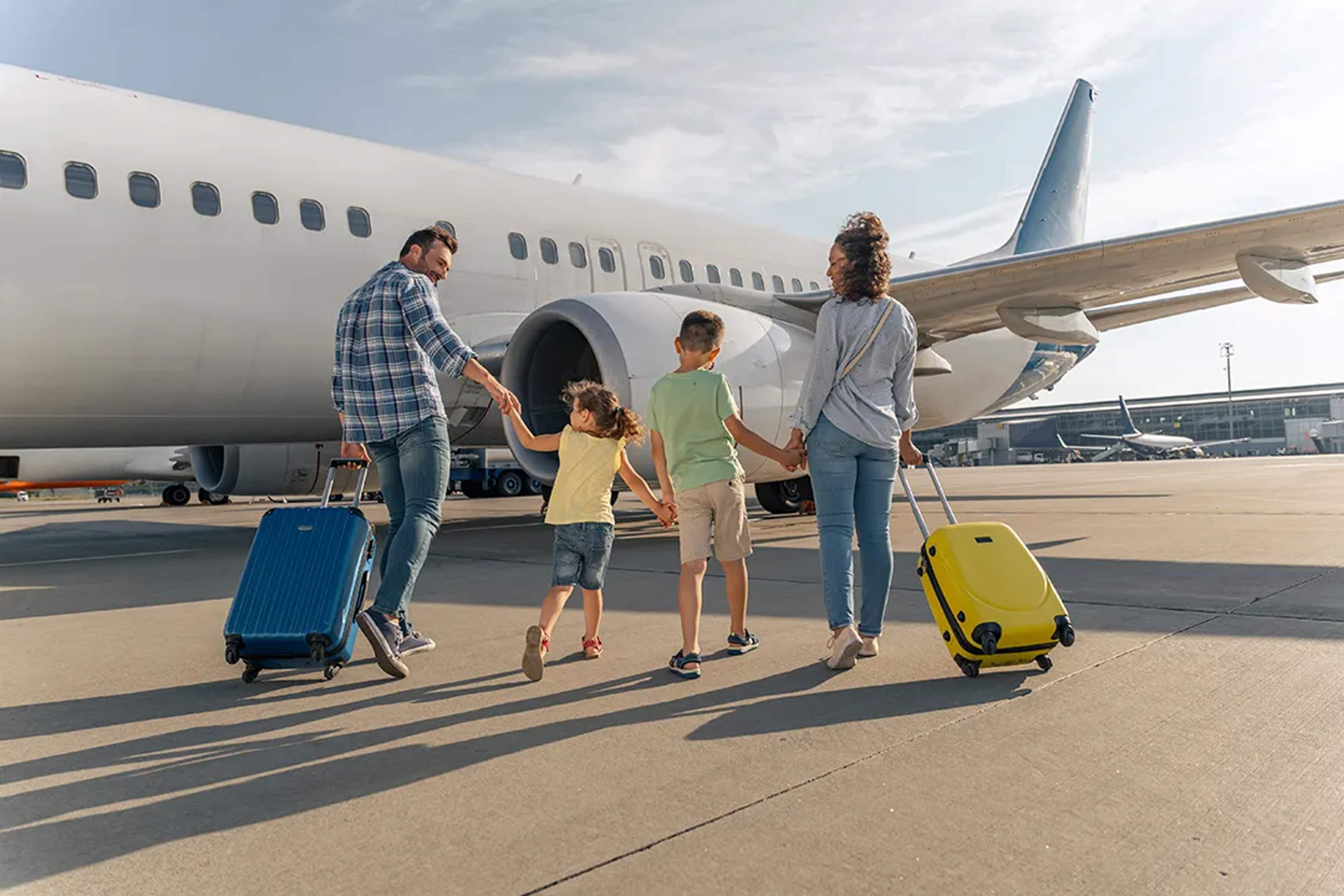 Familia con dos niños camina hacia un avión, cada uno llevando una maleta colorida.