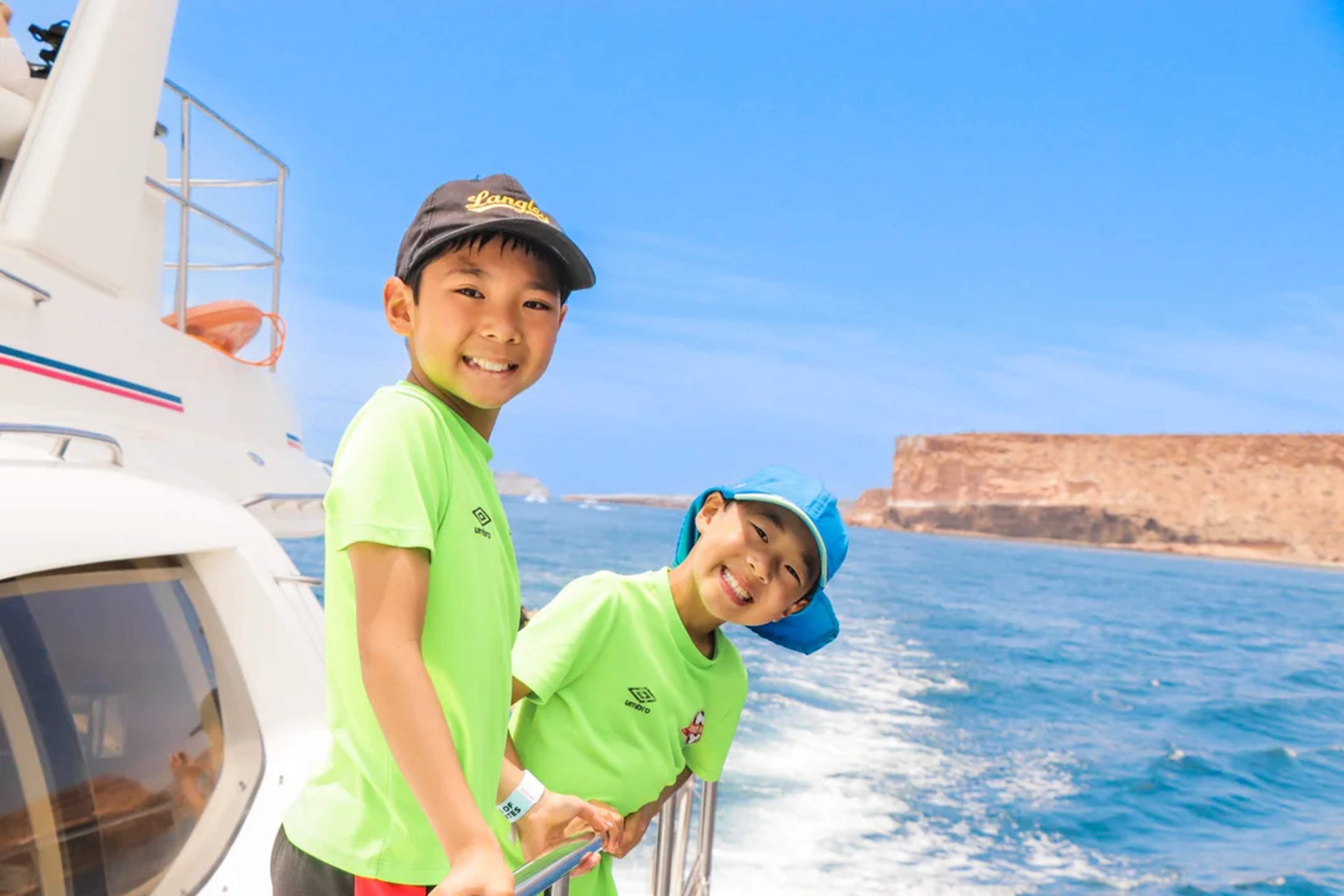Dos niños sonrientes disfrutan un paseo en bote con vistas al mar y acantilados al fondo.