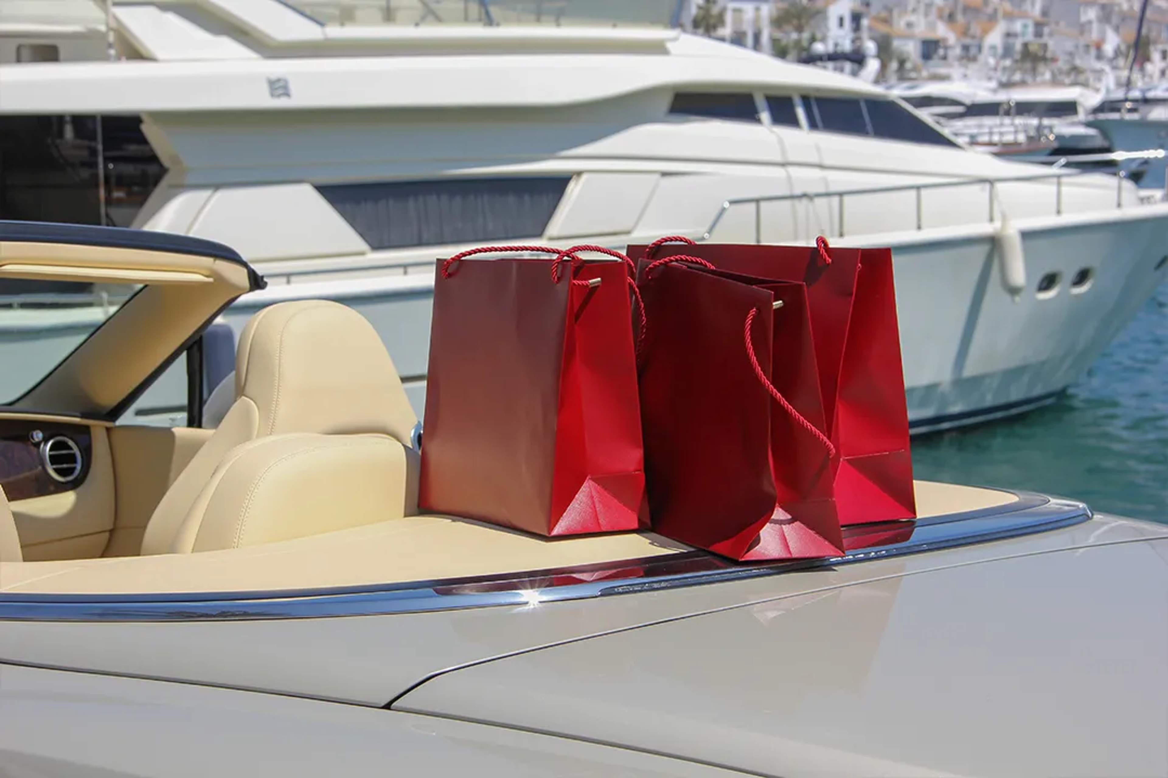 Bolsas rojas de compras sobre un convertible de lujo junto a yates, símbolo de compras exclusivas.