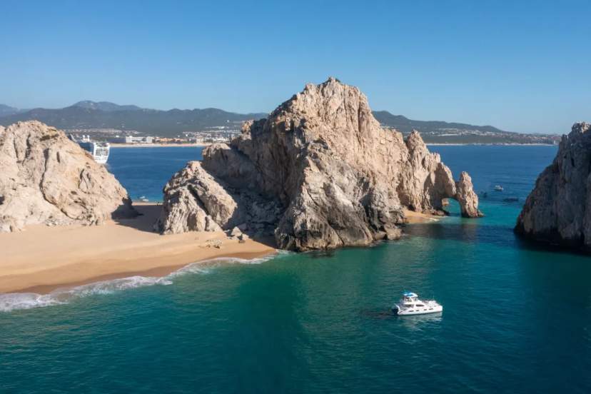 Dramatic rock formations frame a turquoise bay with a snorkel boat anchored along the Cabo San Lucas coastline.