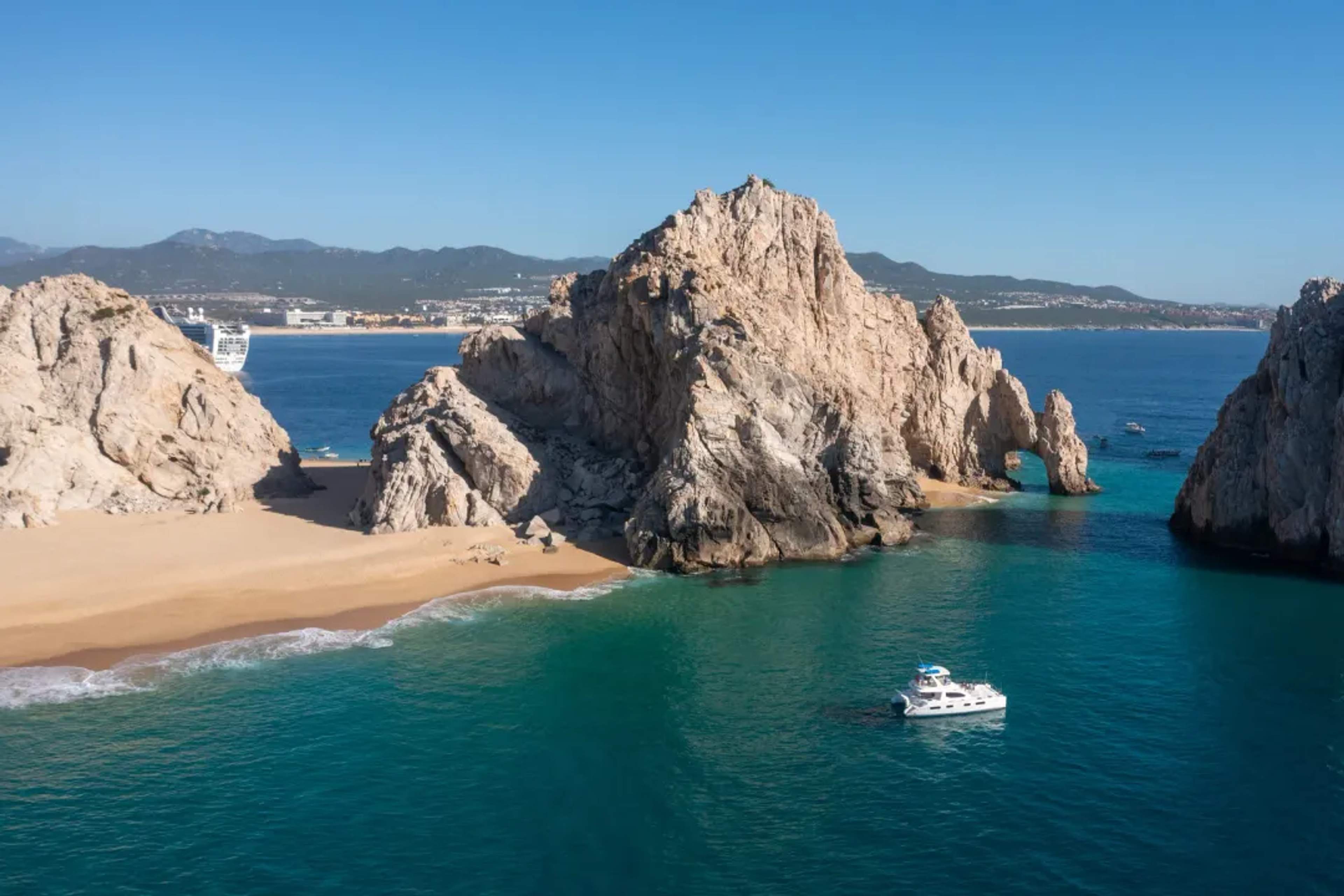 Dramatic rock formations frame a turquoise bay with a snorkel boat anchored along the Cabo San Lucas coastline.