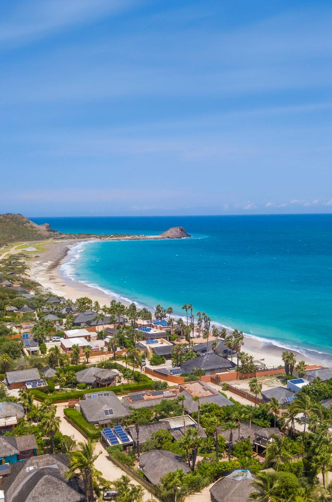 Aerial view of a coastal village with houses and greenery along a sandy beach and turquoise ocean.