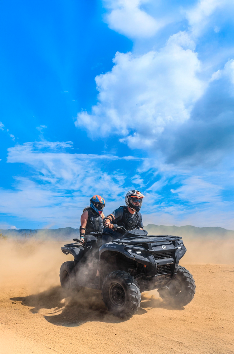 Two riders on an ATV speed through a dusty trail under a bright blue sky, surrounded by scenic mountains and clouds.