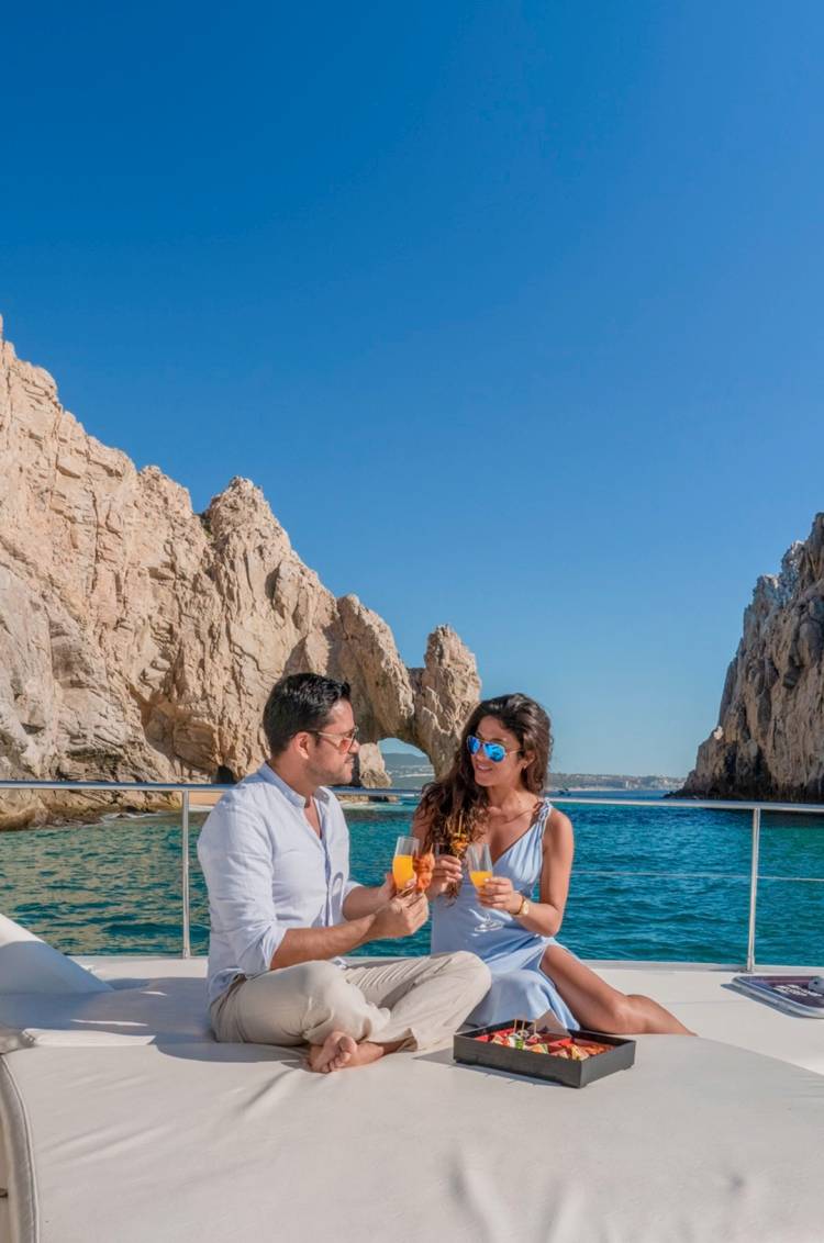 Una pareja disfruta de bebidas y aperitivos en un barco de lujo con vista al famoso Arco de Cabo bajo un cielo azul claro.