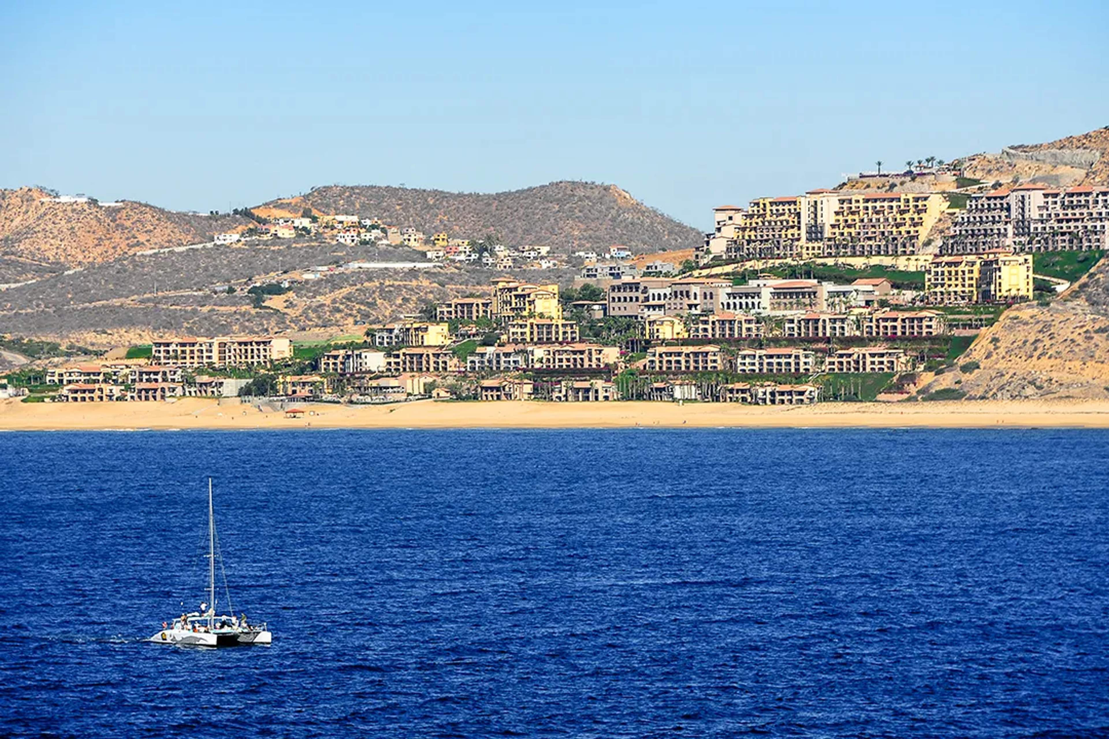 Luxury resorts along Chileno Bay in Los Cabos, with a sailboat cruising deep blue waters and desert hills