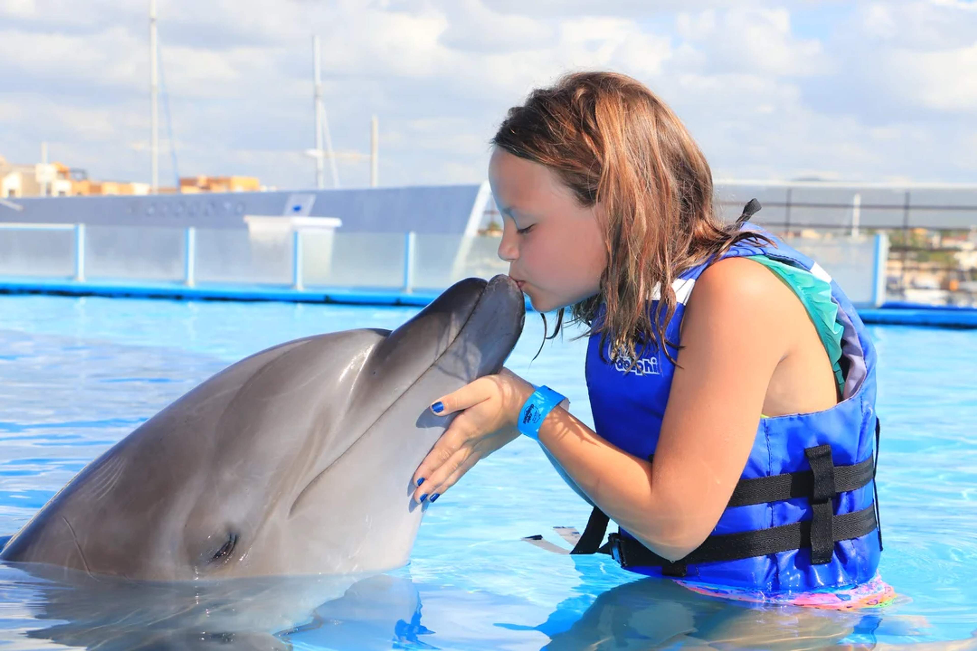 Una niña con chaleco salvavidas comparte un tierno beso con un delfín en un encuentro marino, creando un momento mágico.