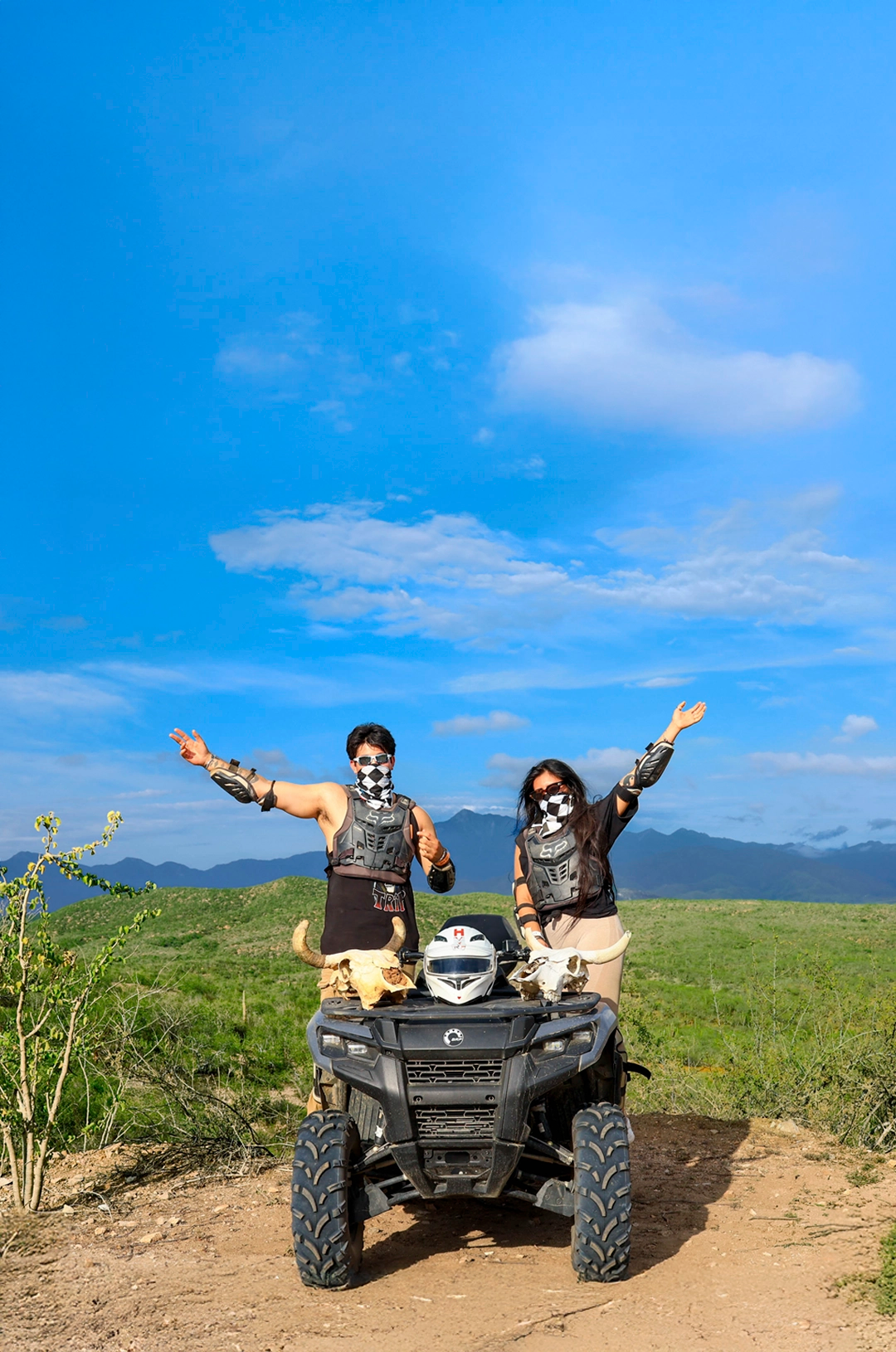 Two people in protective gear pose on an ATV decorated with animal skulls, against a clear blue sky and green mountains.
