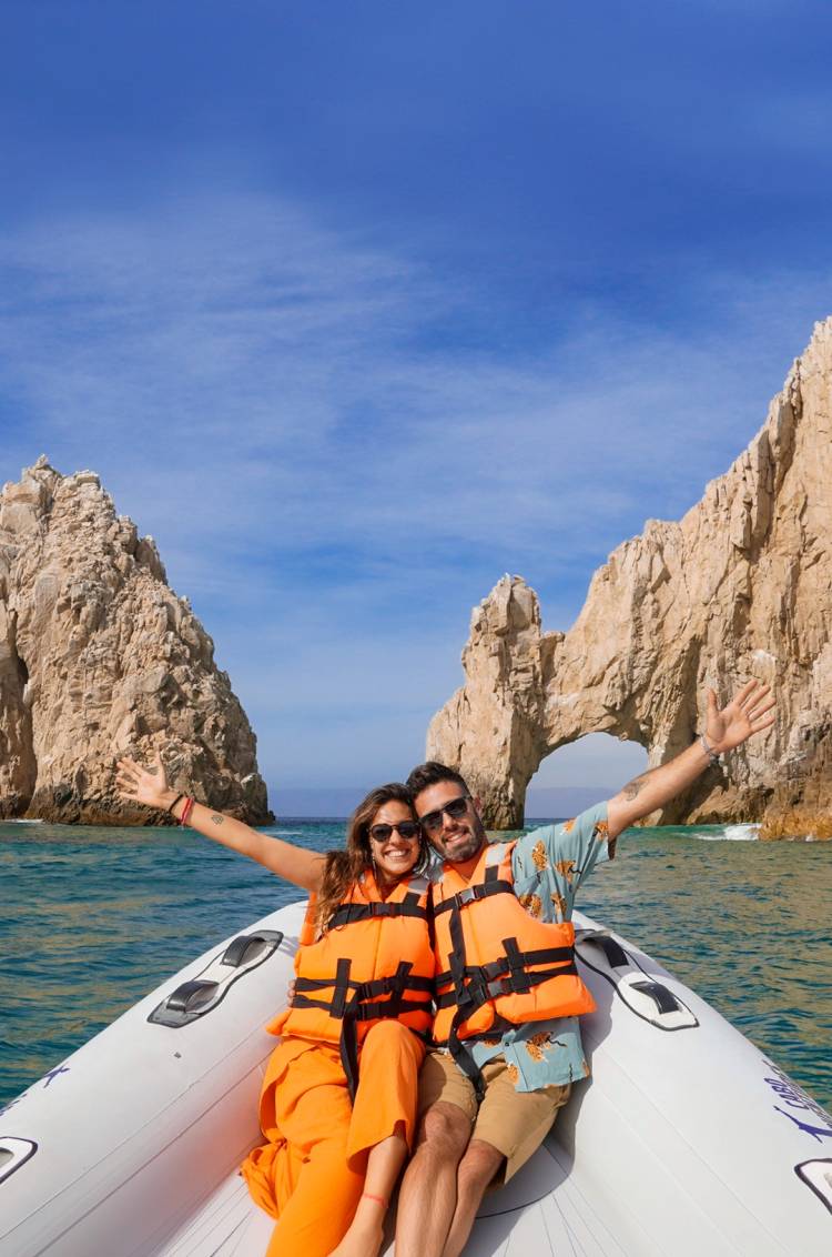 Pareja sonriente con chalecos salvavidas en un bote con una formación rocosa en arco al fondo.