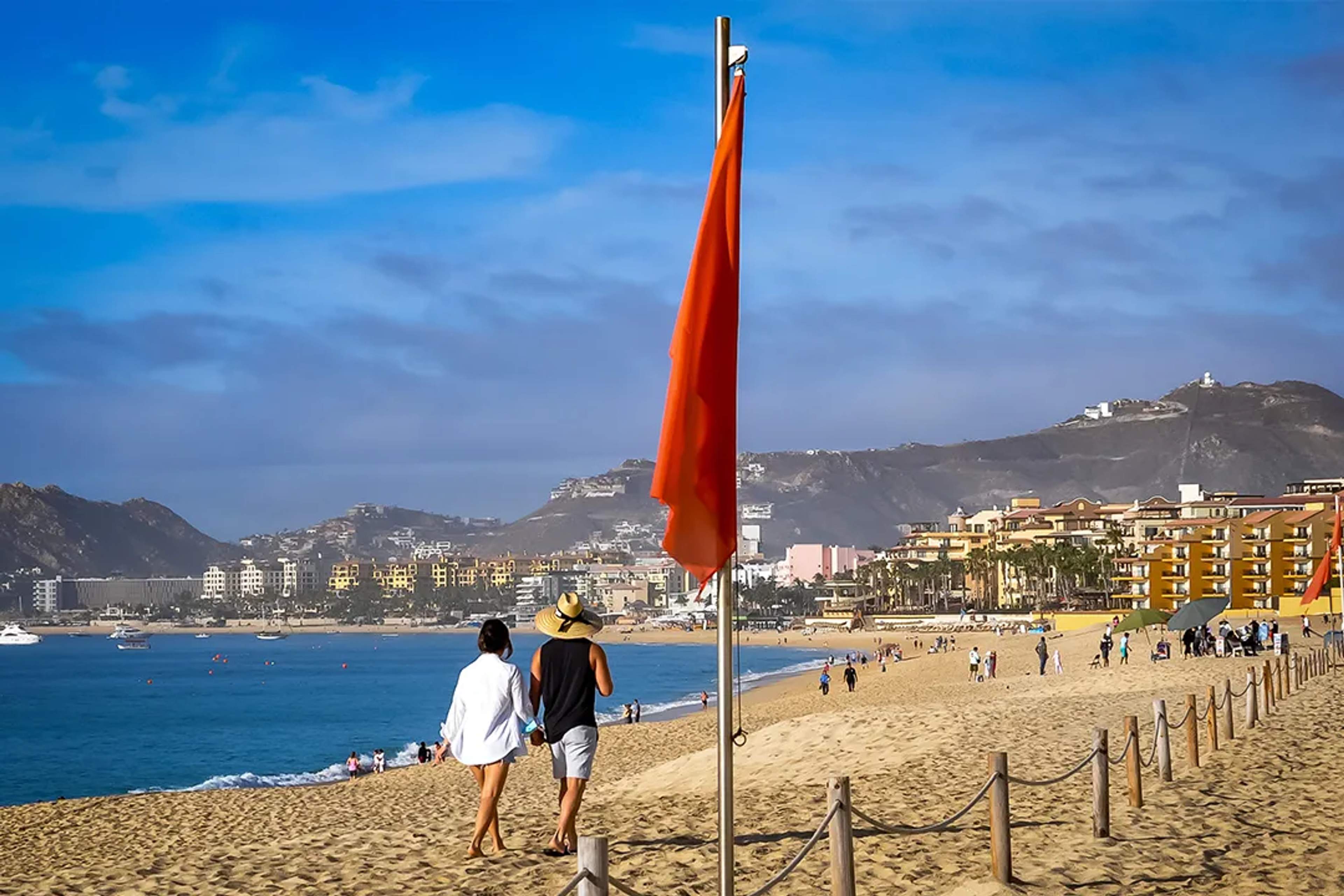Bandera roja indica precaución mientras visitantes caminan por Playa El Médano en Cabo San Lucas.