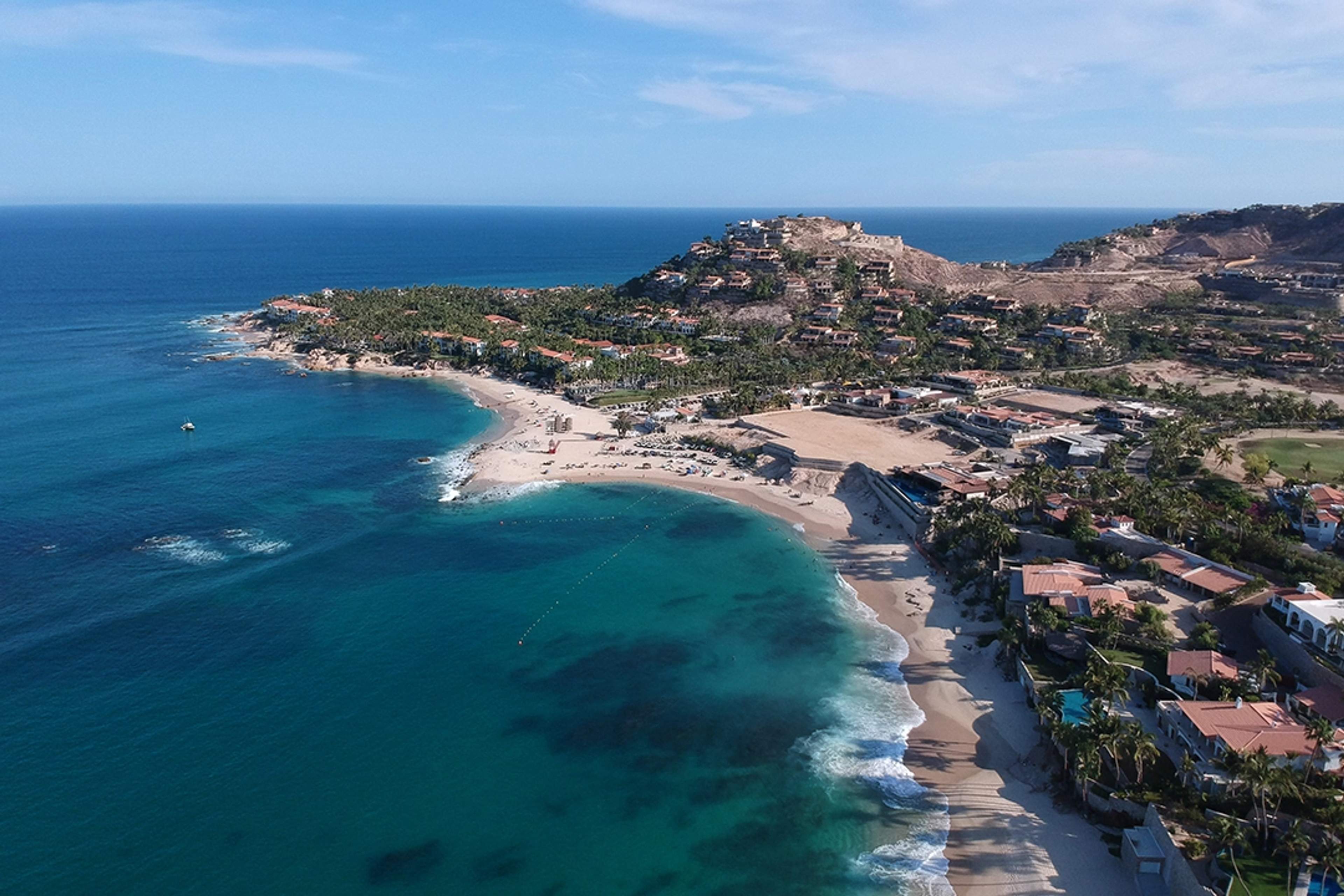 Aerial view of a serene beach surrounded by palm trees and villas, with clear waters in Cabo San Lucas.