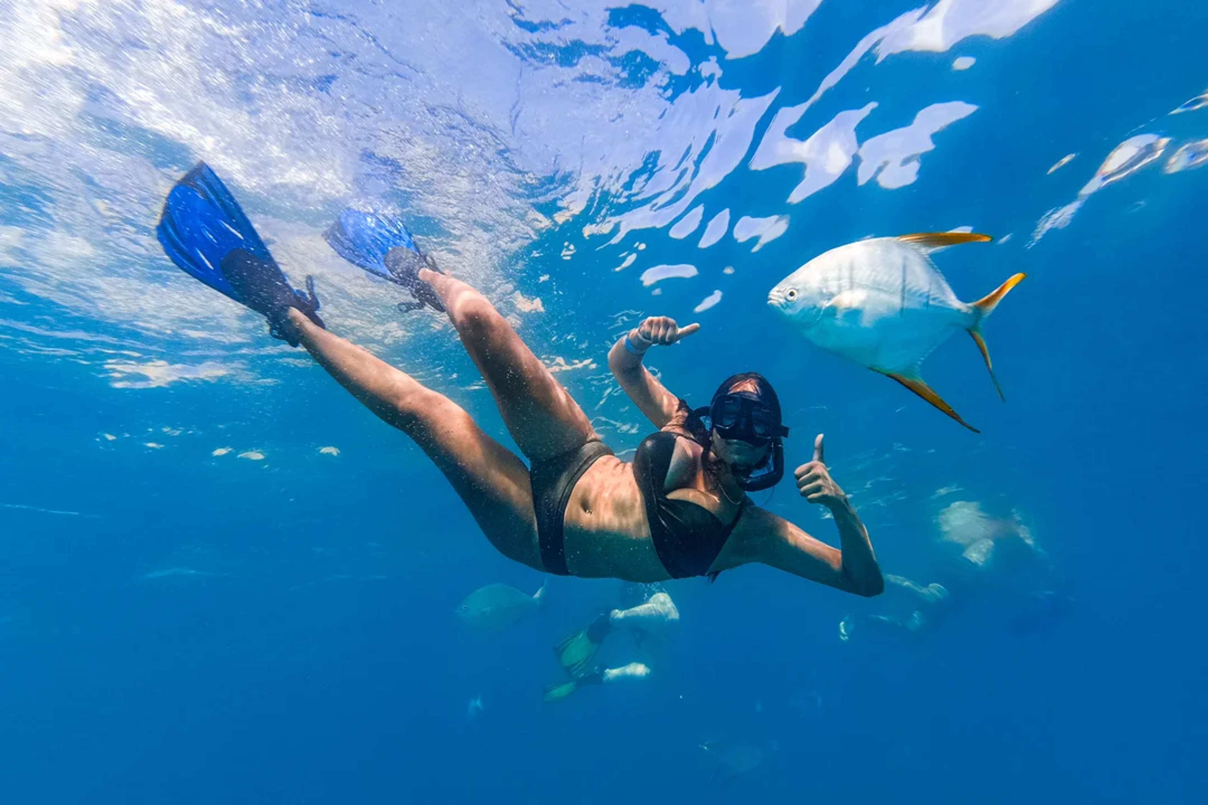 Woman snorkeling with tropical fish, giving thumbs up in clear blue water.