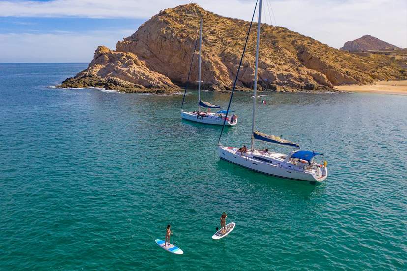 Two women paddleboarding near yachts in a calm bay with rocky hills in the background.