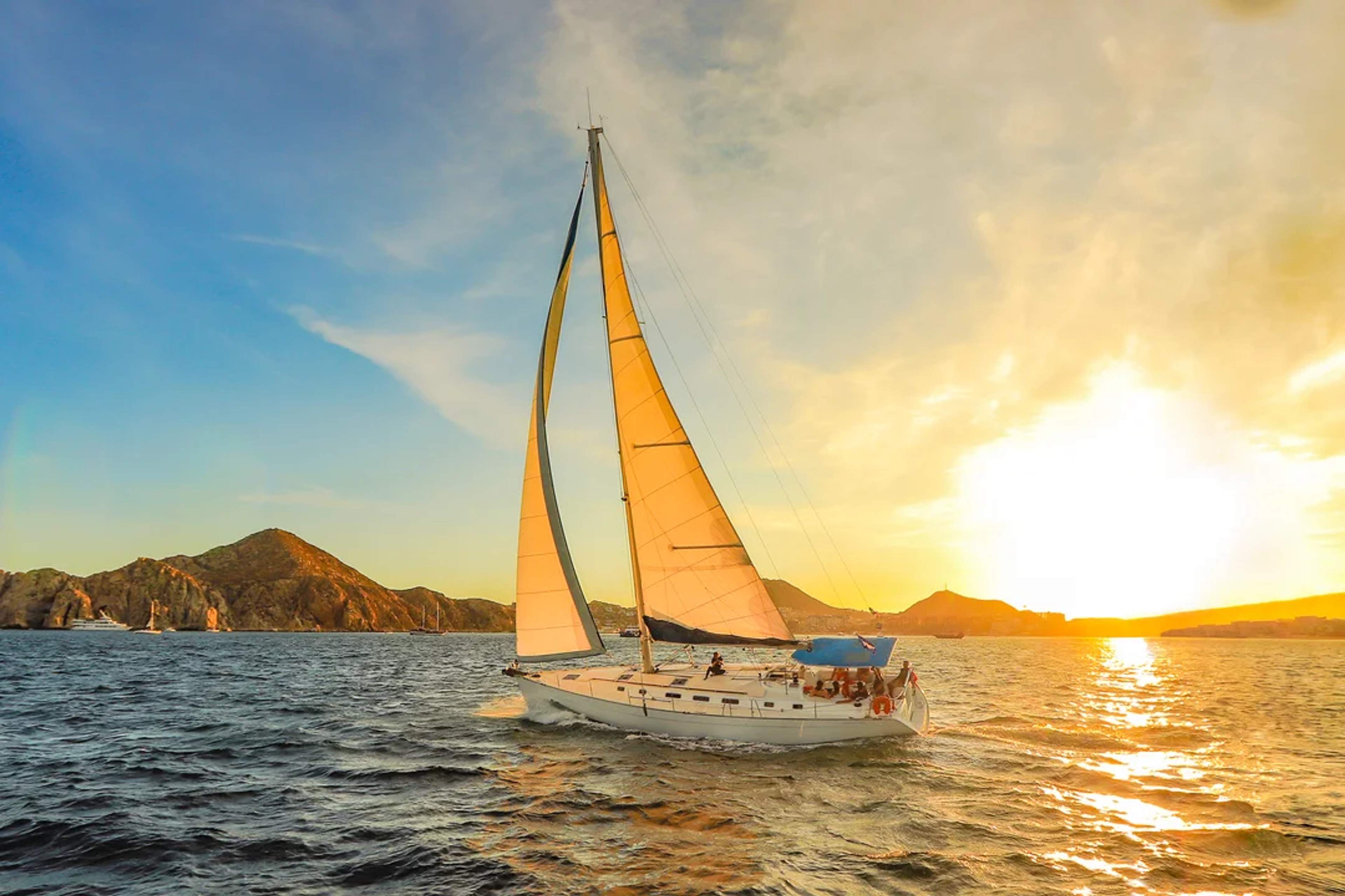 Sailboat gliding at sunset near rocky shores, with golden skies and peaceful ocean views.