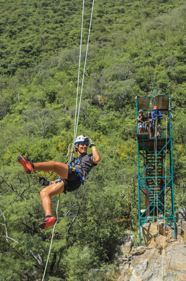 A person zip-lining through a forested area with a tall platform in the background, wearing a helmet and harness.