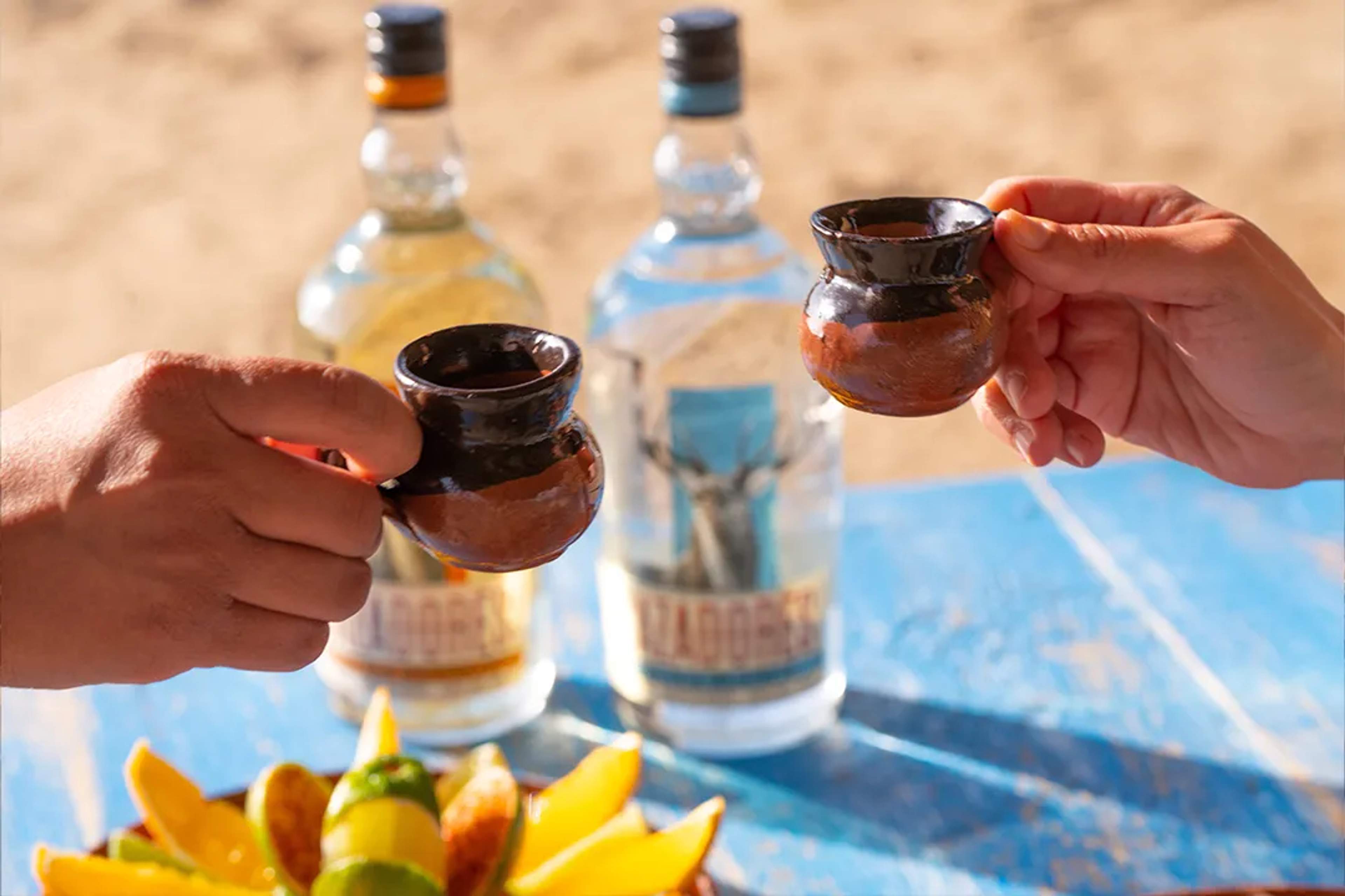Two people toast with clay cups of tequila by the beach, with fruit and bottles in the background.