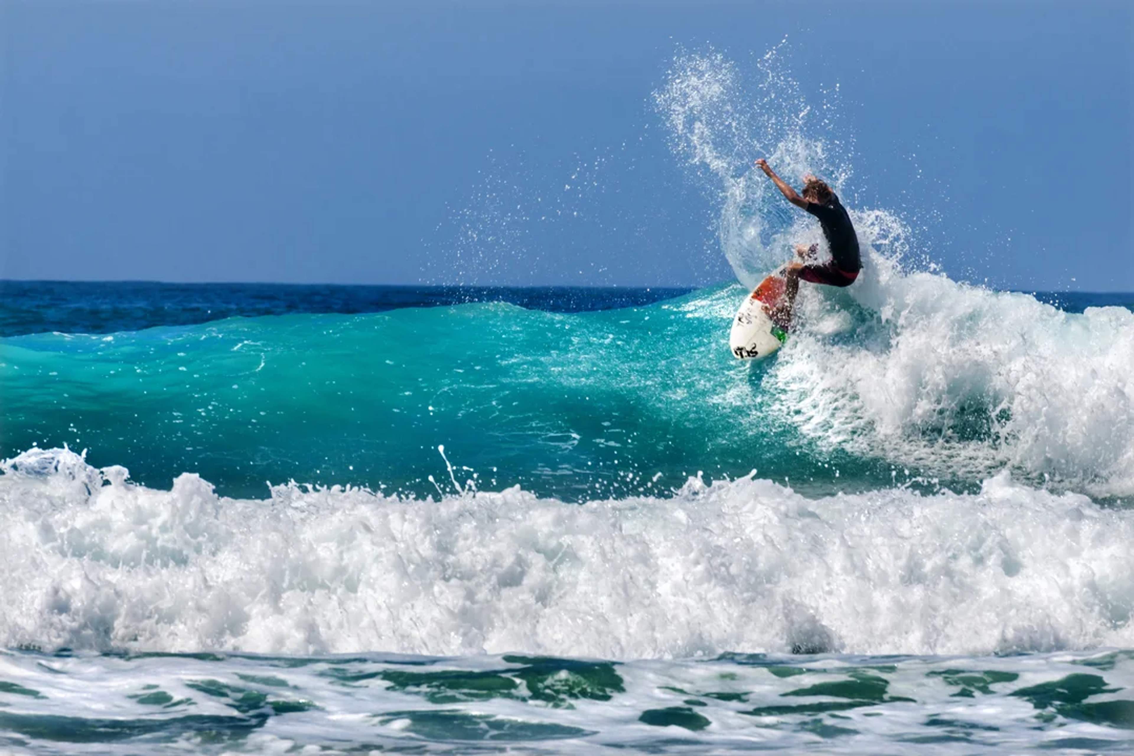 Un surfista domina una ola turquesa, surcando el agua mientras el océano salpica bajo un cielo azul brillante.