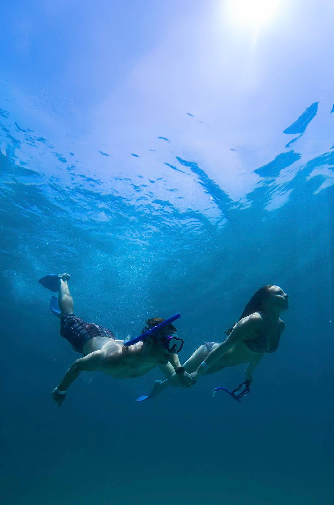 Una pareja hace esnórquel bajo el agua, nadando de la mano en aguas azules, con la luz del sol filtrándose desde arriba.
