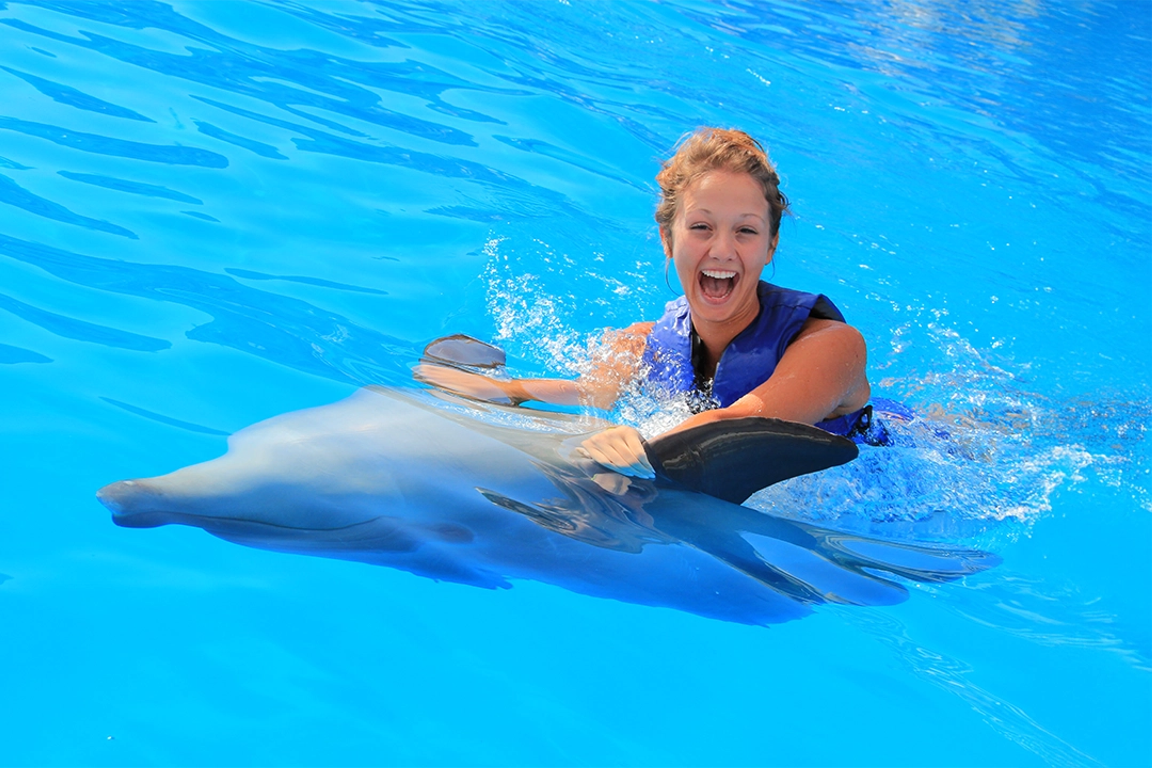 Smiling woman enjoying a ride with a dolphin in clear waters, wearing a life jacket.