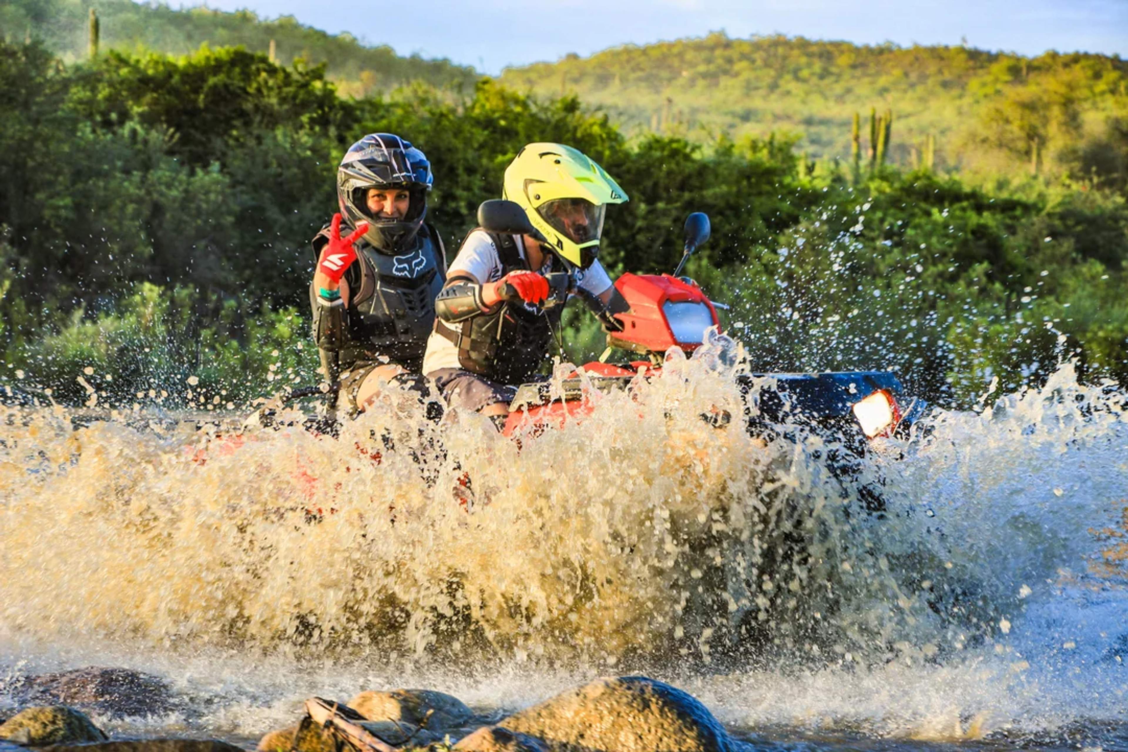 Dos personas cruzan un arroyo en cuatrimoto, entre vegetación desértica y pura adrenalina.